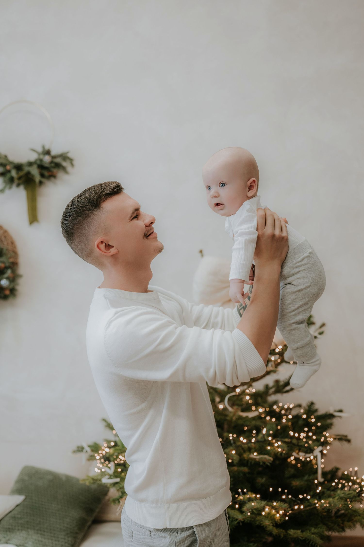 A young father in a white sweater joyfully lifts his baby near a decorated Christmas tree with warm lights.
