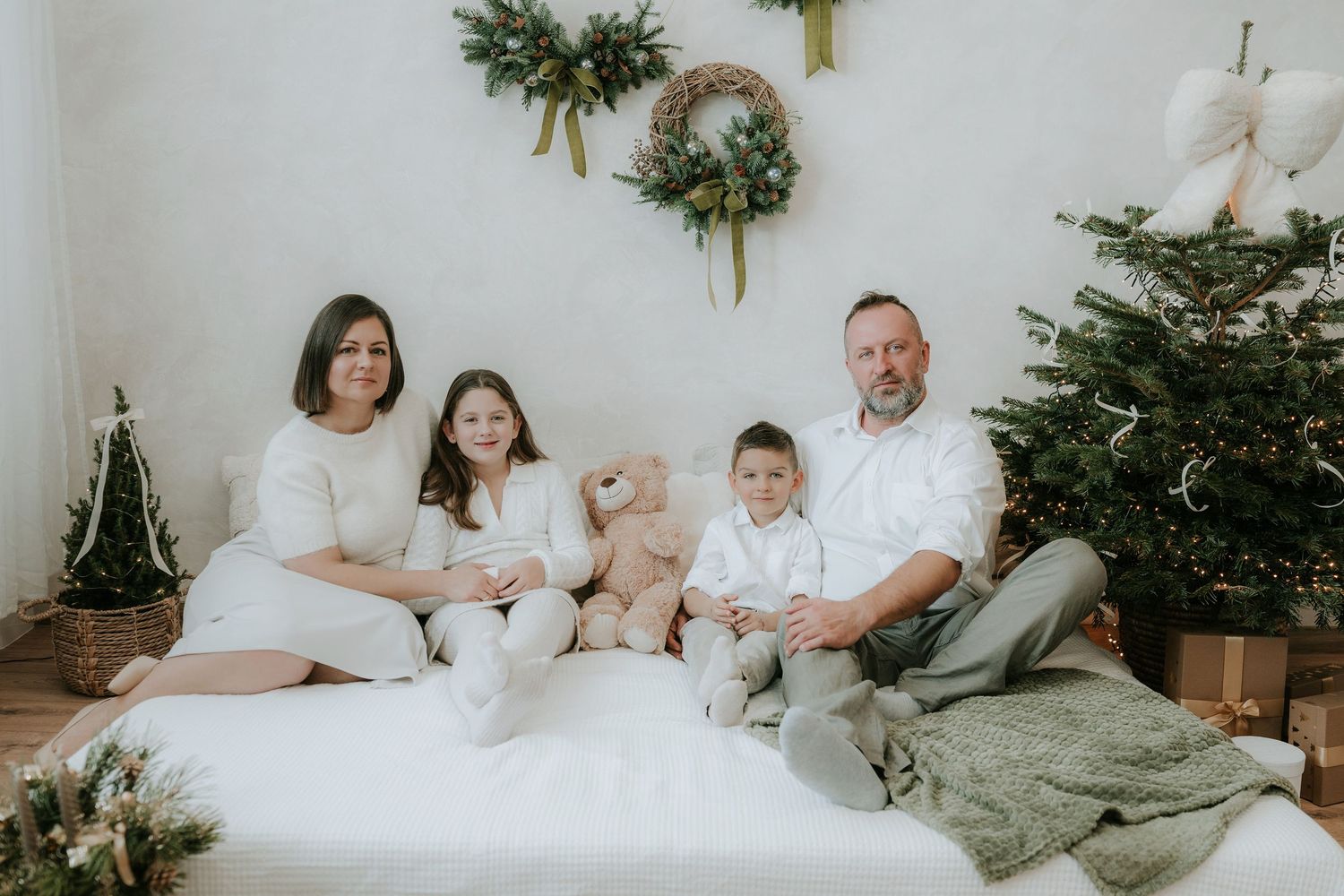 Family of four posing for a Christmas photo on a white bed with a teddy bear and decorated tree.