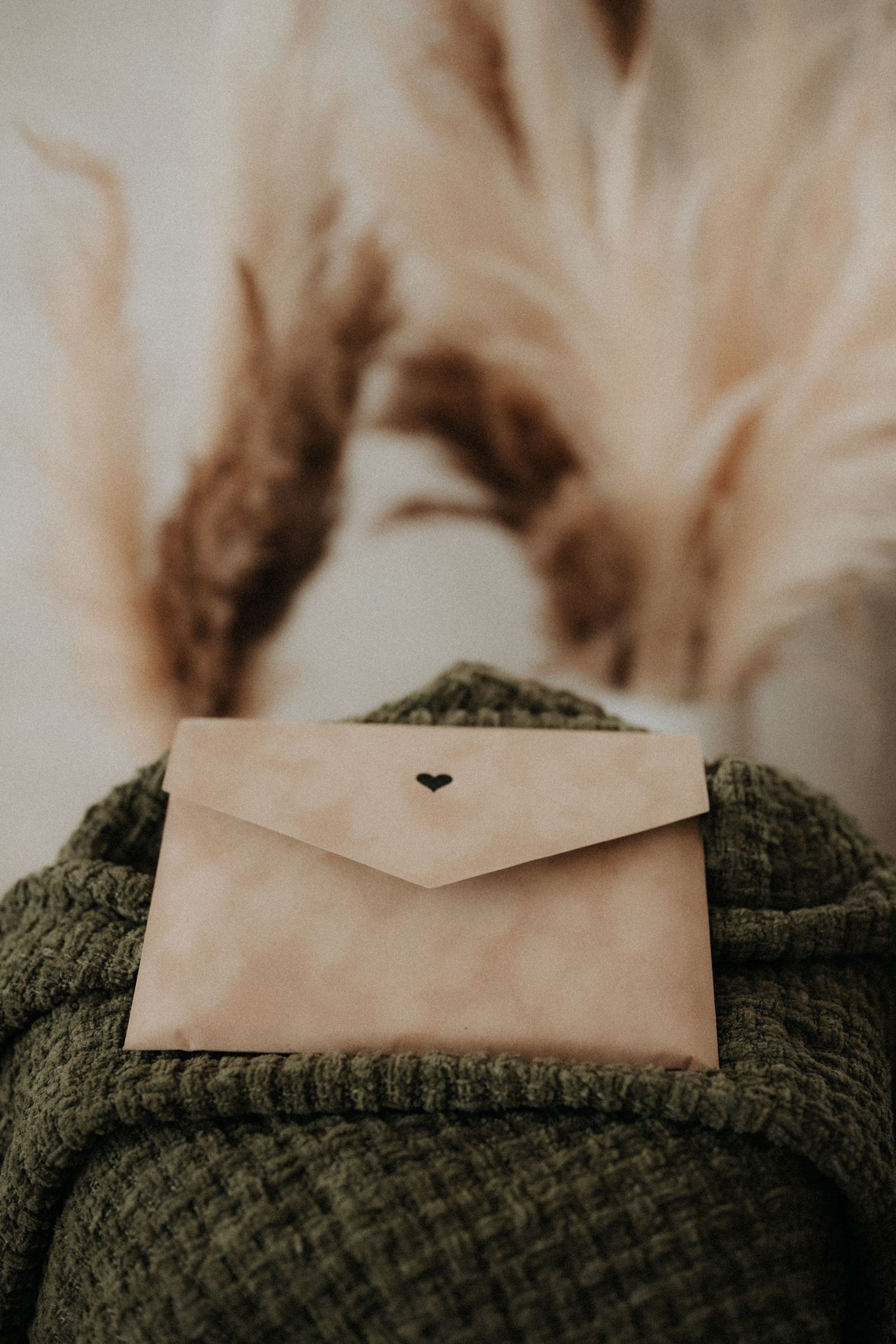 A beige envelope with a heart detail rests on a green knit sweater with pampas grass in the background.