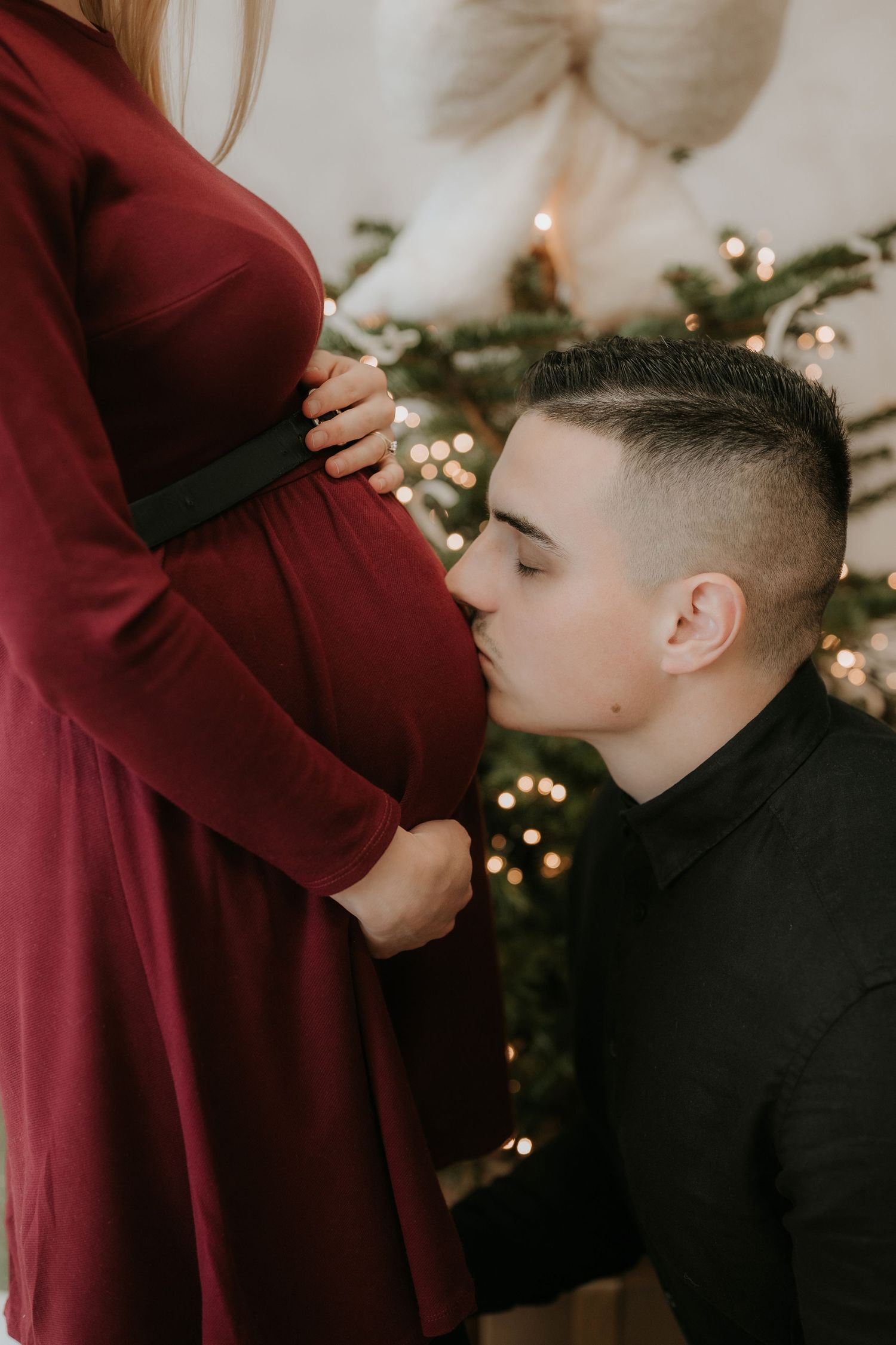 A young man kisses a pregnant woman's belly in front of a decorated Christmas tree, capturing a tender maternity moment.