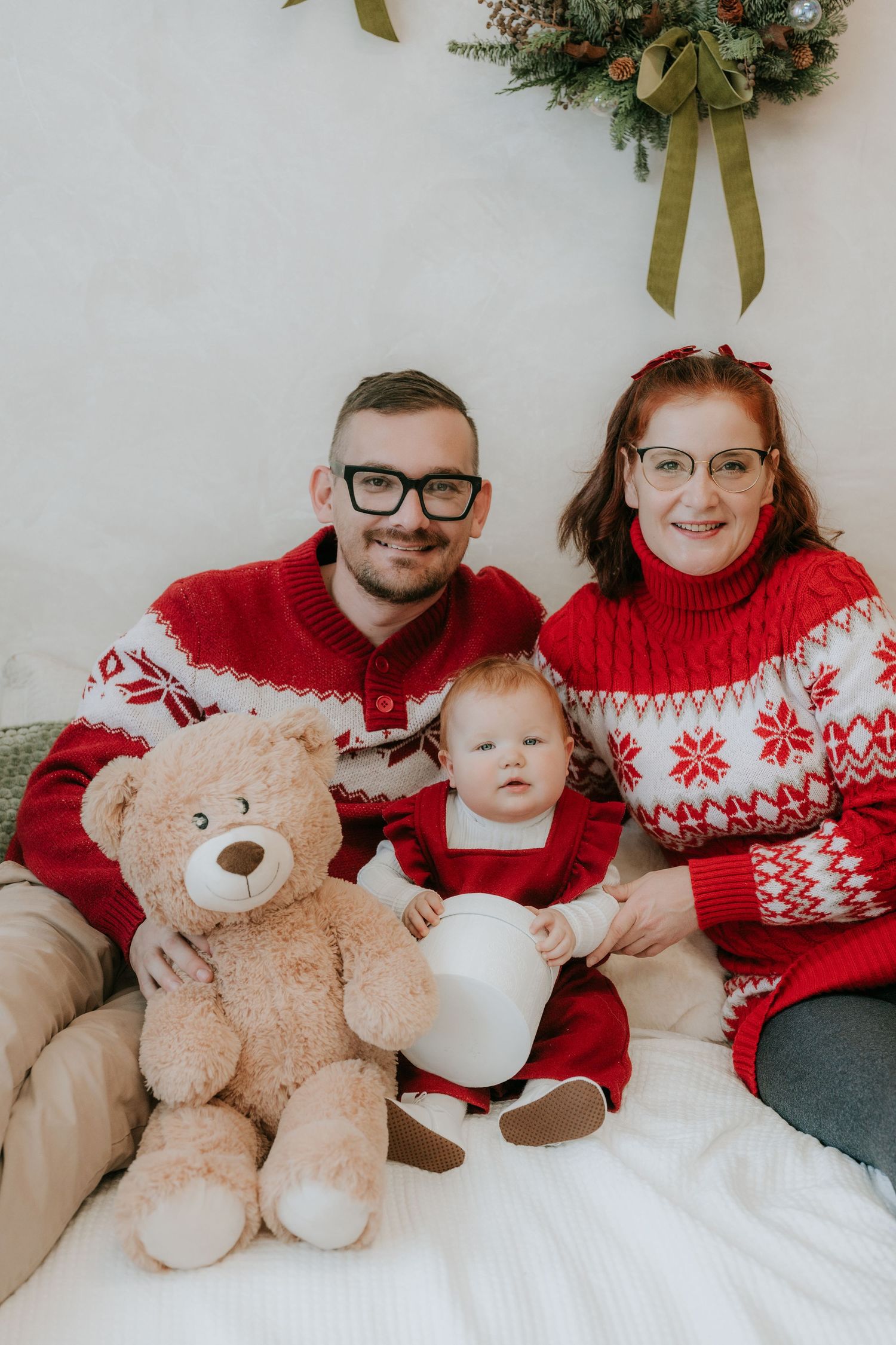Family of three in matching red Nordic sweaters poses with teddy bear for Christmas photo session.