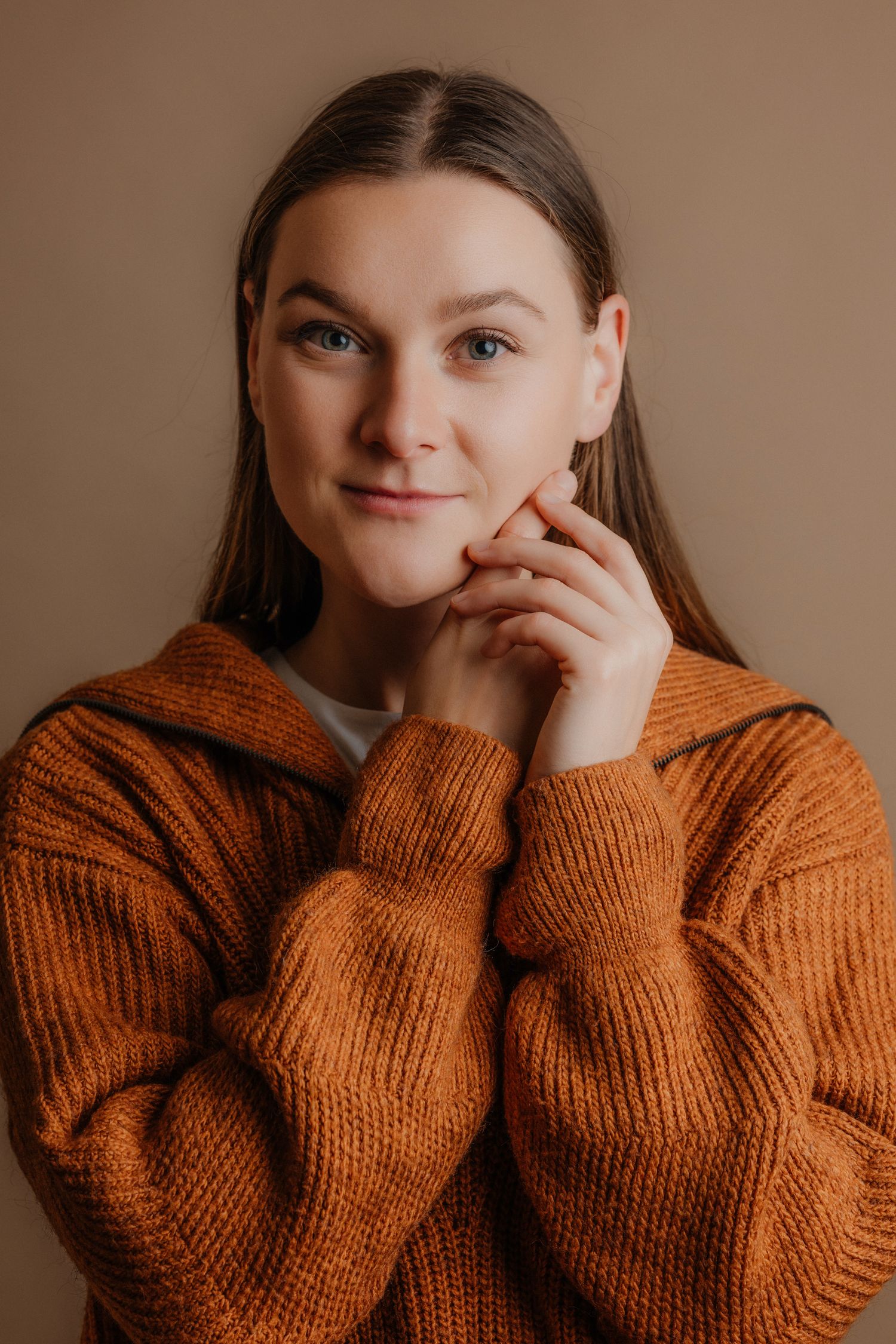 Young woman in orange knit sweater posing thoughtfully with hand near chin against beige background.