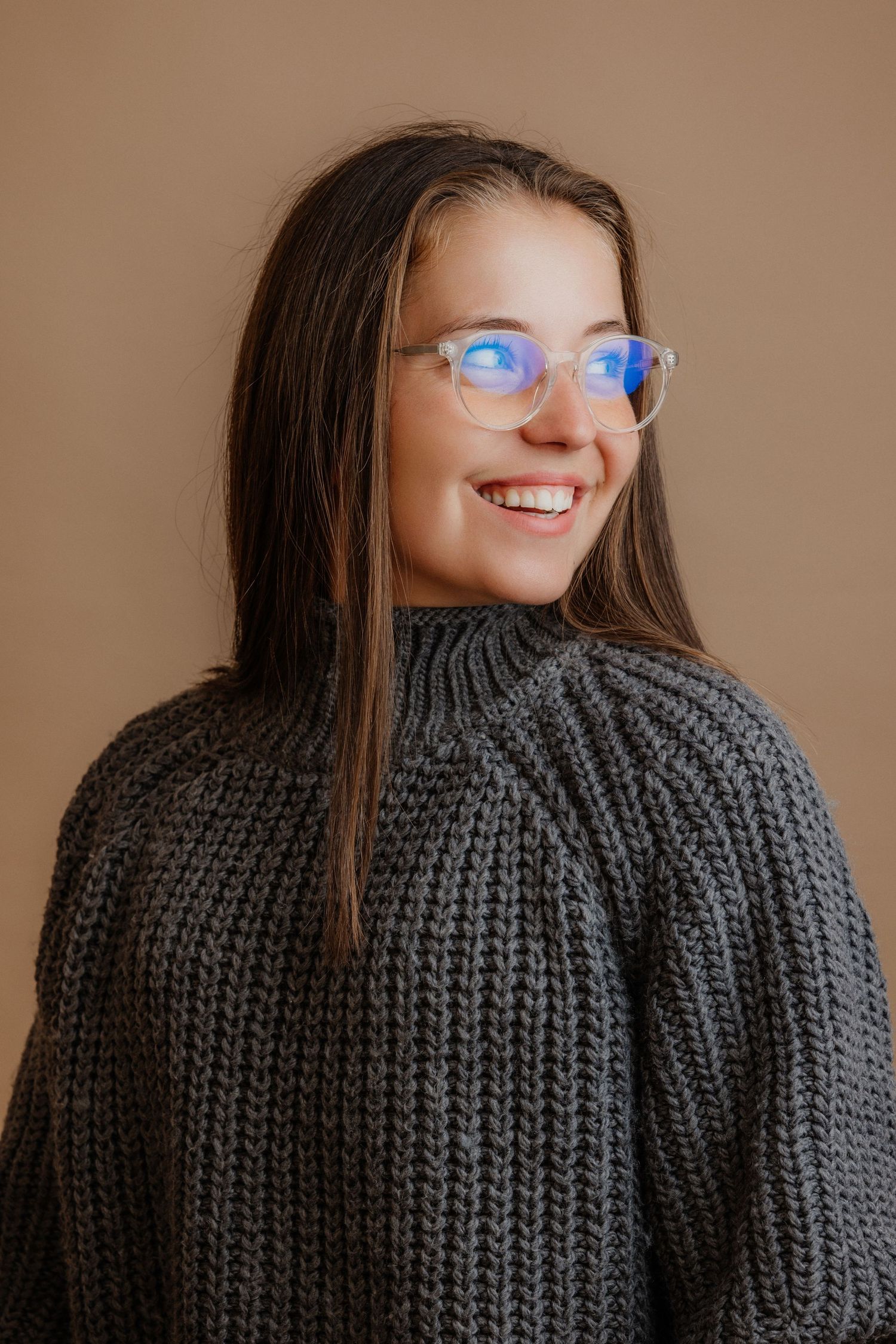 Smiling young woman with long brown hair wearing clear glasses and a chunky knit gray sweater against a beige background.