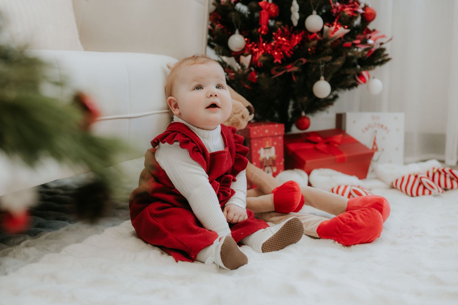 A baby girl in a red dress sits on a white fur rug beside a decorated Christmas tree with gifts.