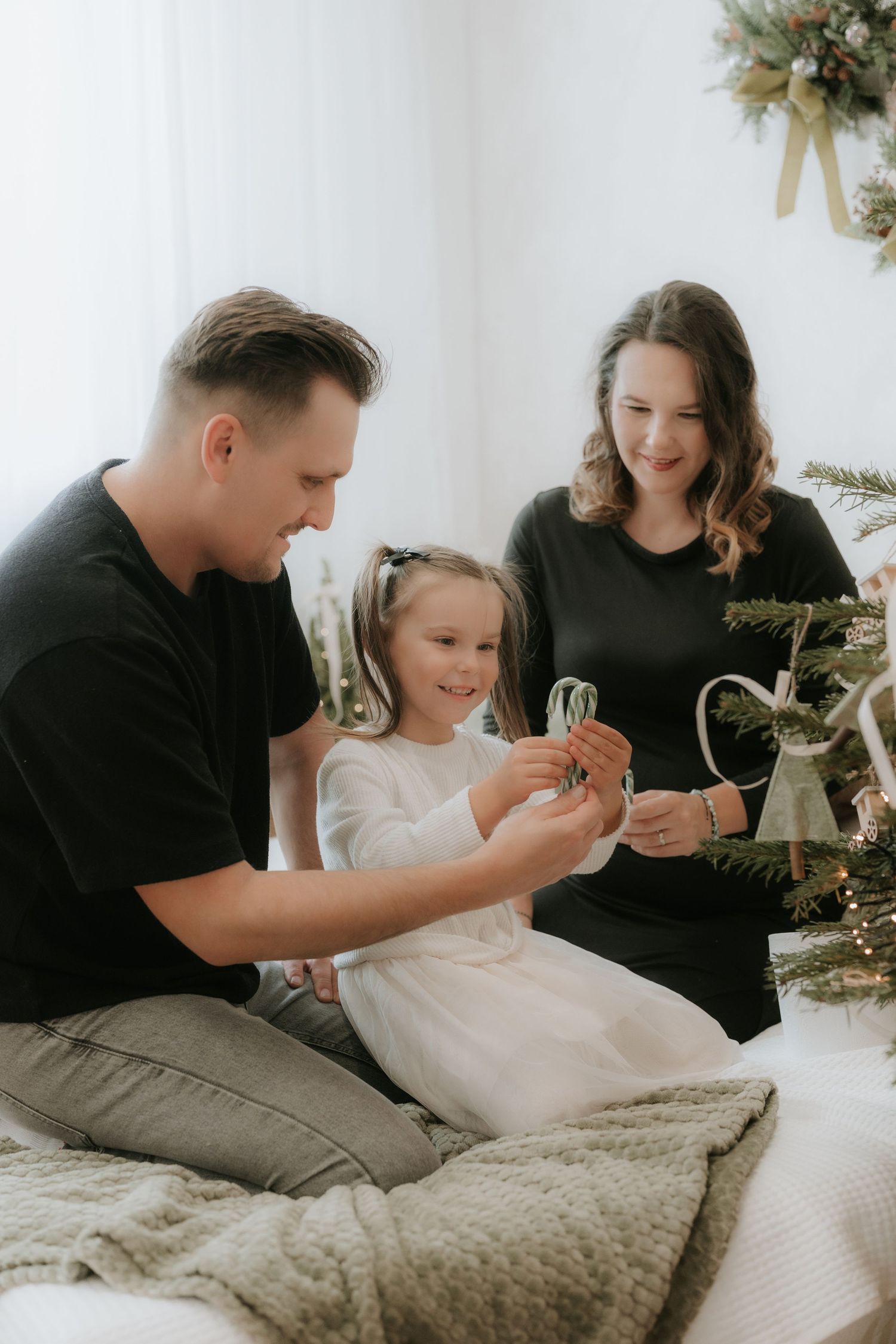 Family decorating Christmas tree together, parents in black shirts with young daughter in white dress holding holiday ornaments.