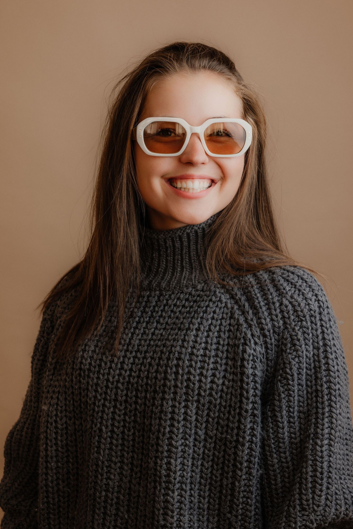 Smiling young woman wearing white rectangular sunglasses and a chunky gray knit turtleneck sweater against a beige background.