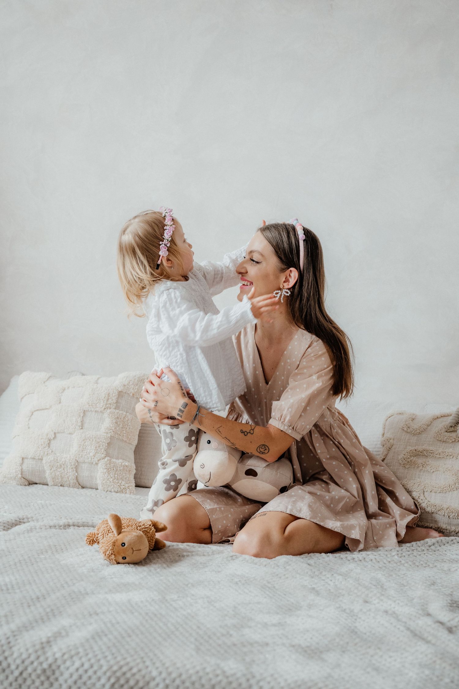 A mother sits on a bed holding her toddler daughter, both smiling, with stuffed animals nearby in a soft, neutral-toned room.