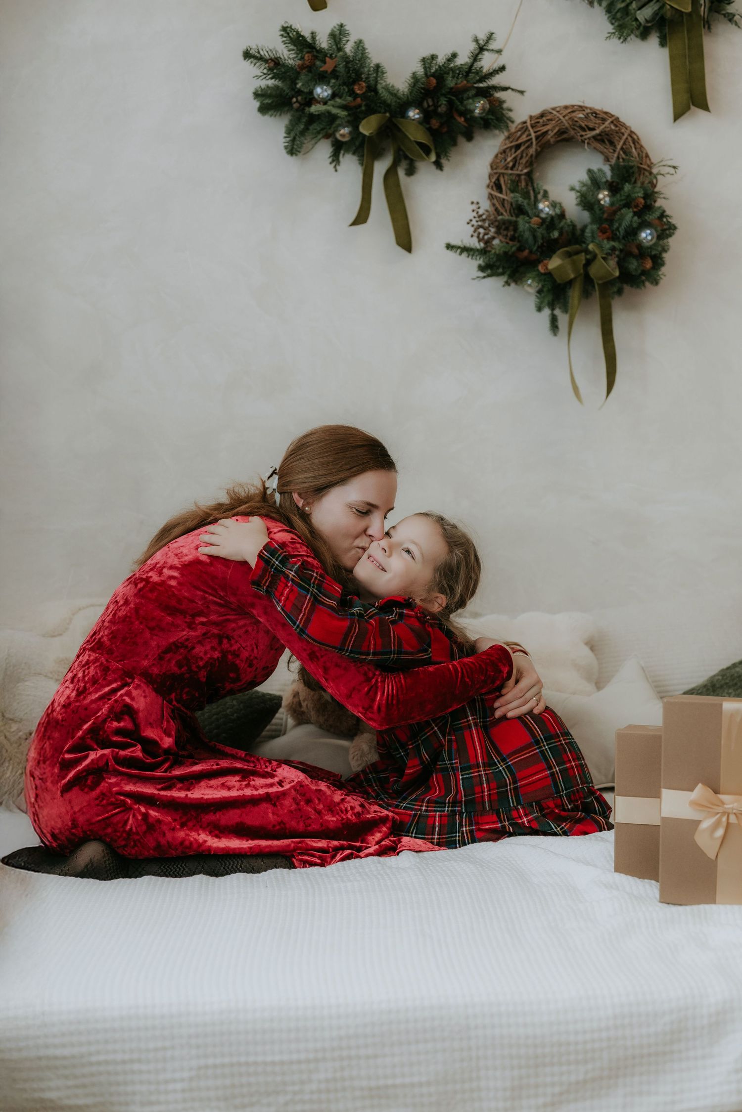Mother and daughter in red Christmas outfits share a warm hug on a bed with holiday wreaths in background.
