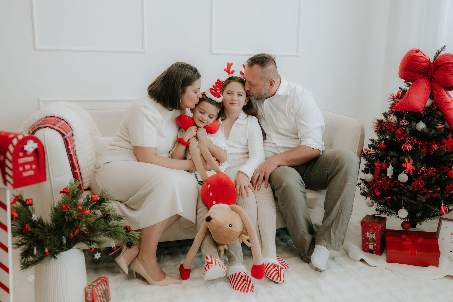 Family in white outfits celebrating Christmas on a sofa with a decorated tree and festive gifts nearby.