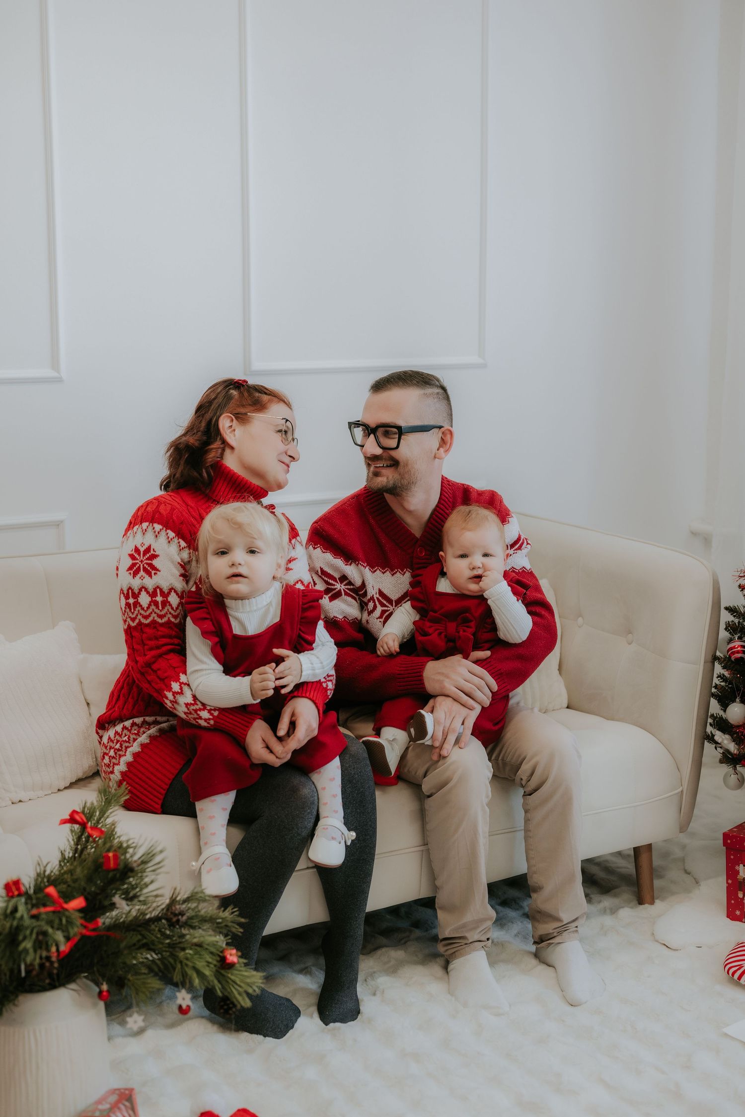 Family of four in matching red Nordic sweaters sitting on a white sofa with Christmas decorations.