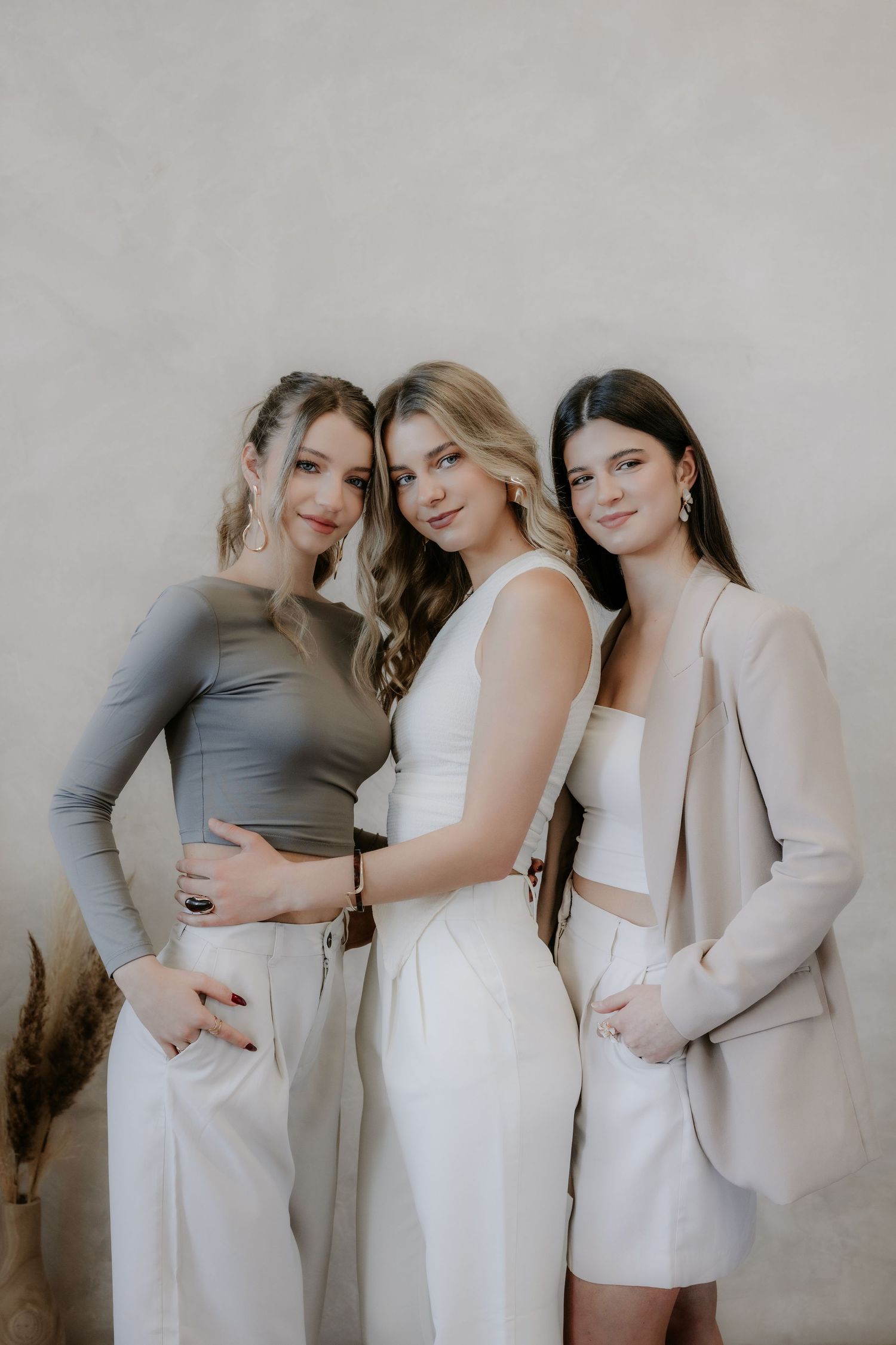 Three young women pose together in neutral tones, wearing stylish outfits of gray, white, and beige against a light wall.