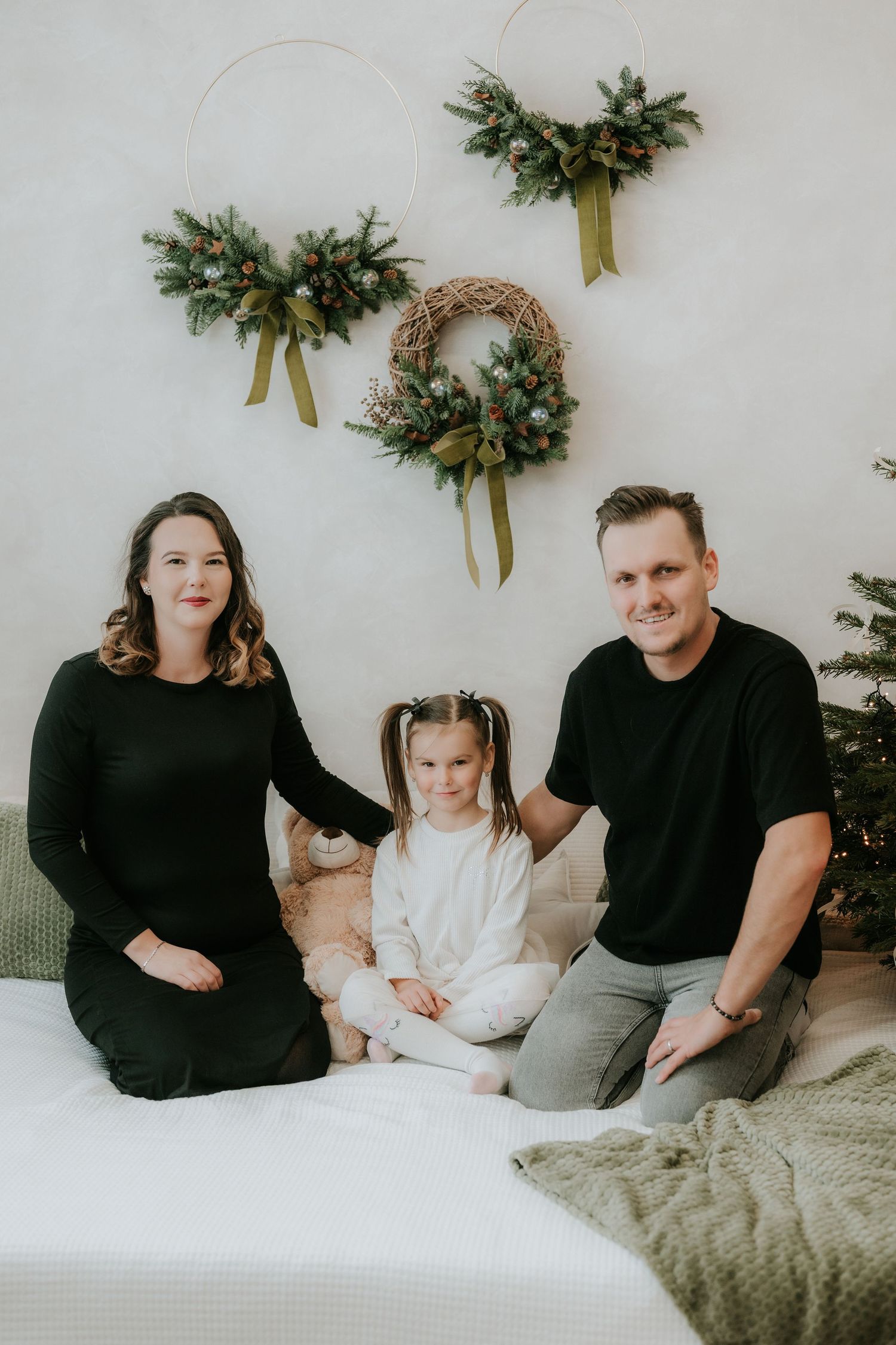 Family of three poses on a bed with Christmas wreaths on the wall behind them in a festive holiday photo.