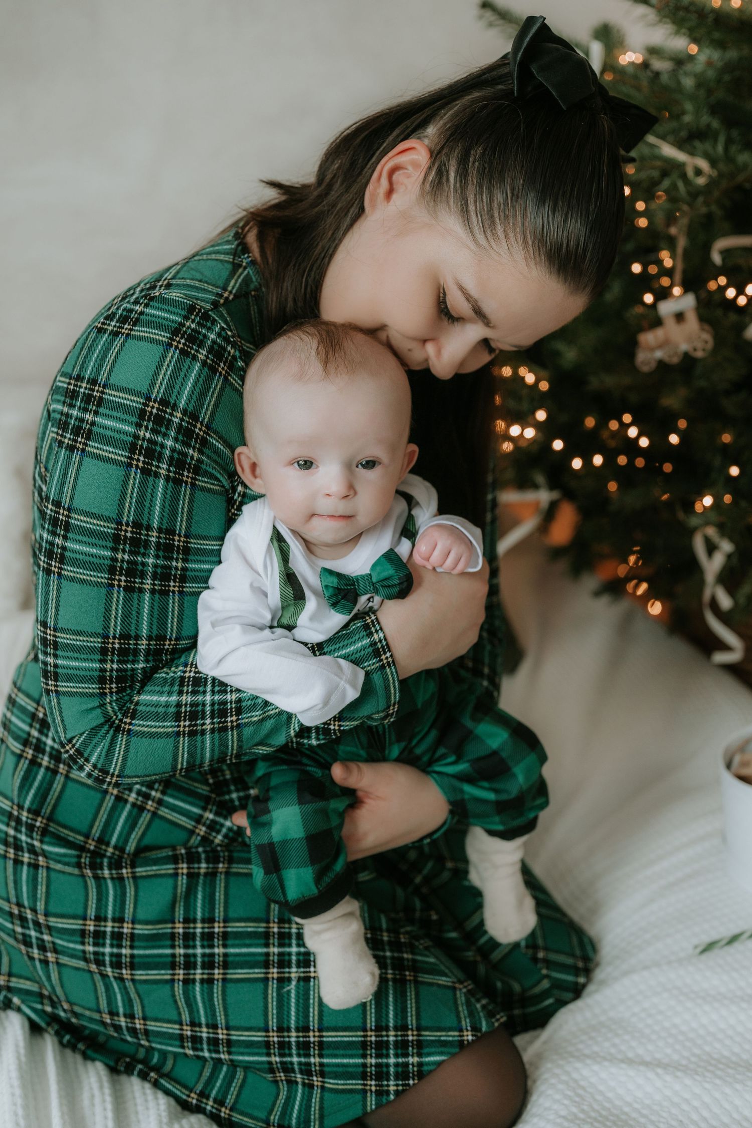 Mother in green plaid dress tenderly kissing baby in matching outfit near Christmas tree.