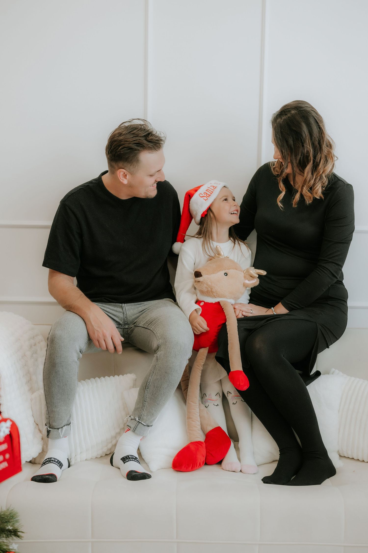 Family Christmas photo with dad, daughter in Santa hat holding reindeer toy, and pregnant mom sitting together.
