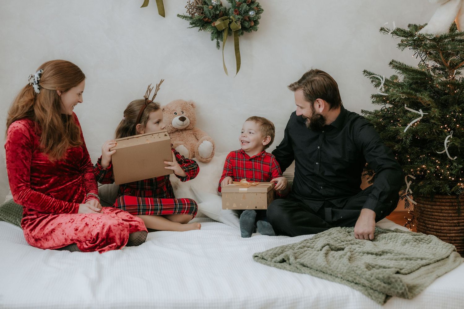 Family of four exchanging Christmas gifts on white bed near decorated tree, children in festive plaid pajamas with teddy bear.
