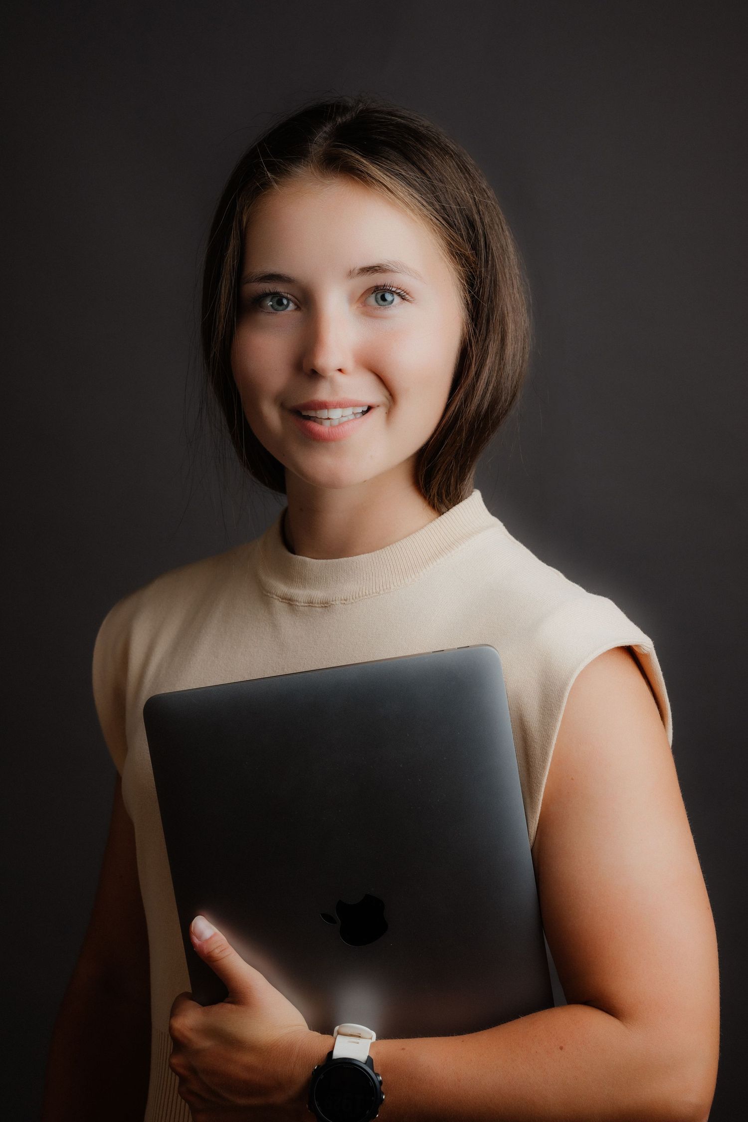 Young woman smiling, holding a MacBook laptop, wearing a beige sleeveless top and watch, against a dark background.