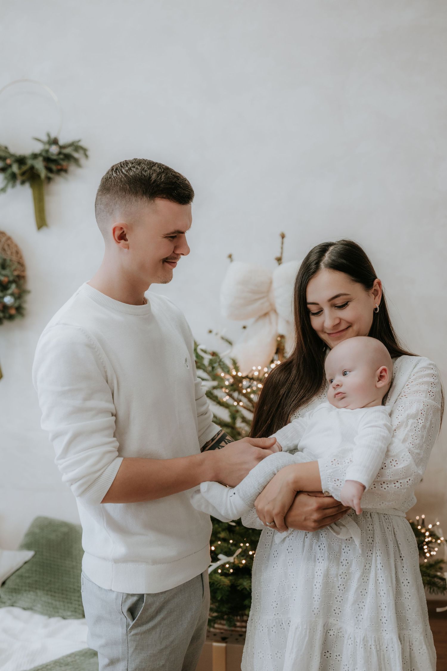 Young family with newborn baby posing near Christmas tree decorations in matching white outfits.