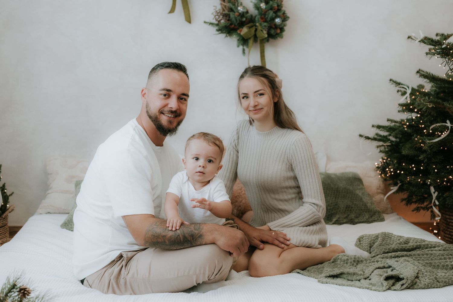 Young family with baby sitting on white bed near Christmas tree in cozy holiday photo session.