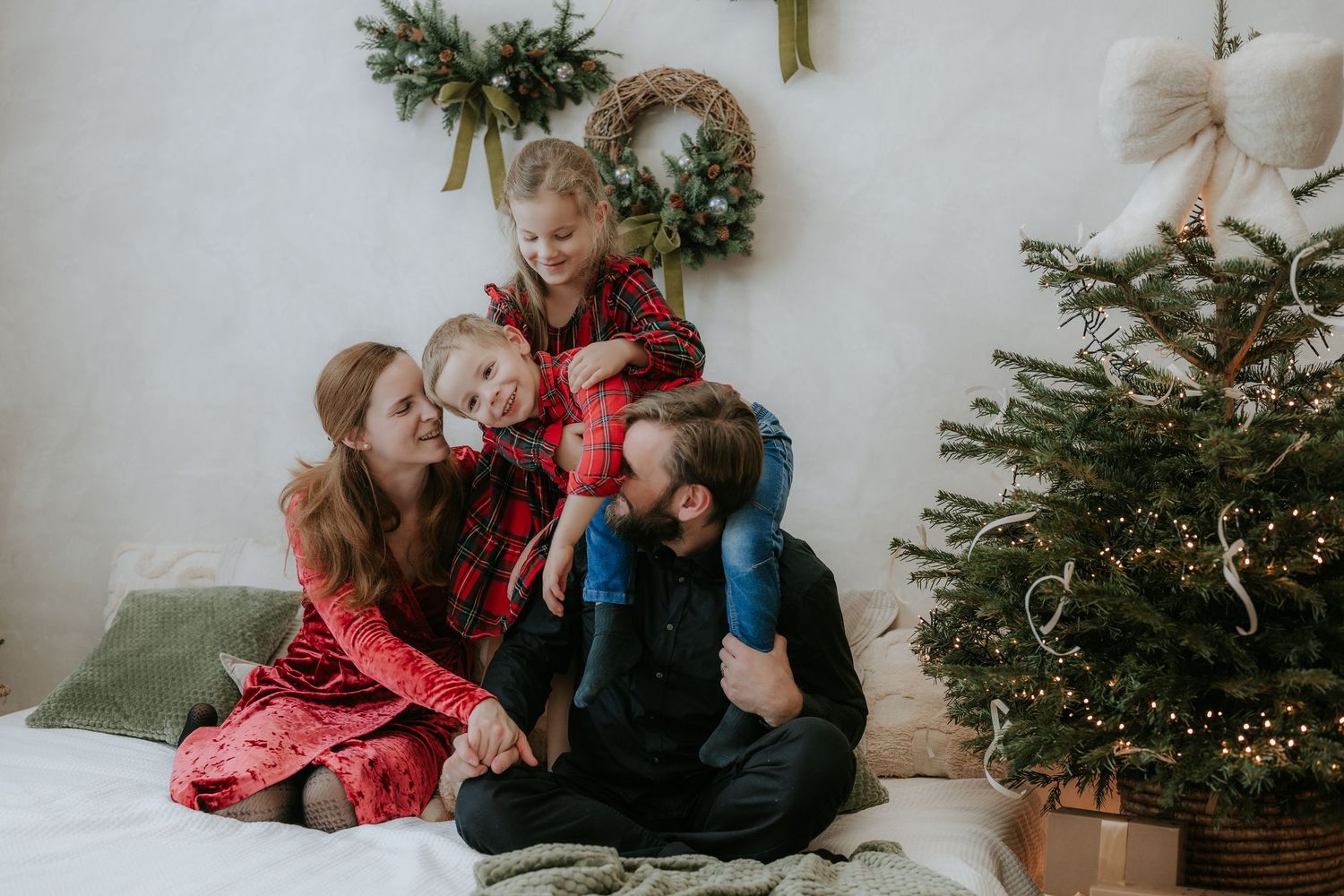 Family in matching red plaid pajamas laughing together near a decorated Christmas tree with wreath backdrop.