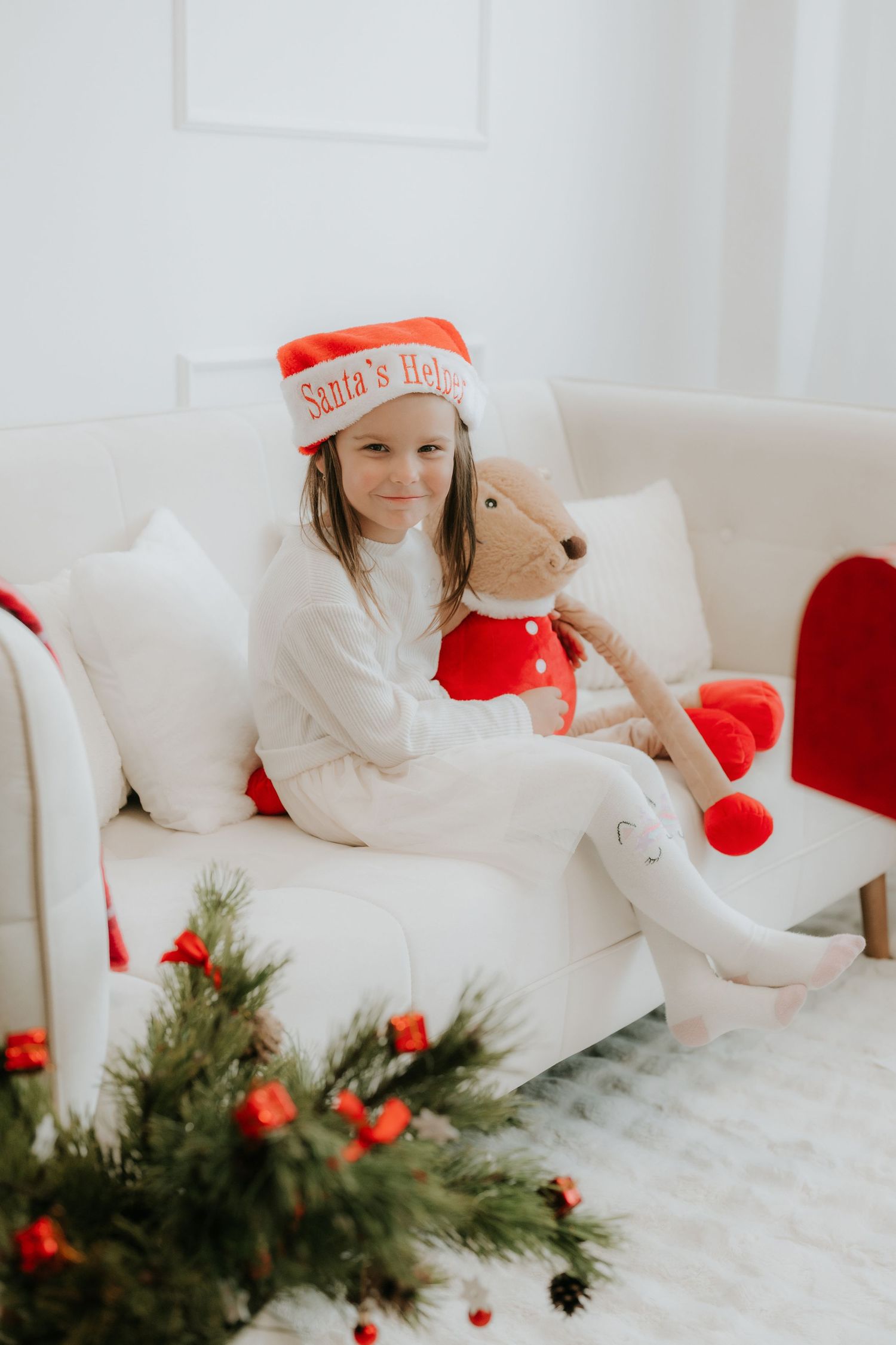 Young girl in Santa's Helper hat sits on white sofa hugging a teddy bear with Christmas decorations nearby.