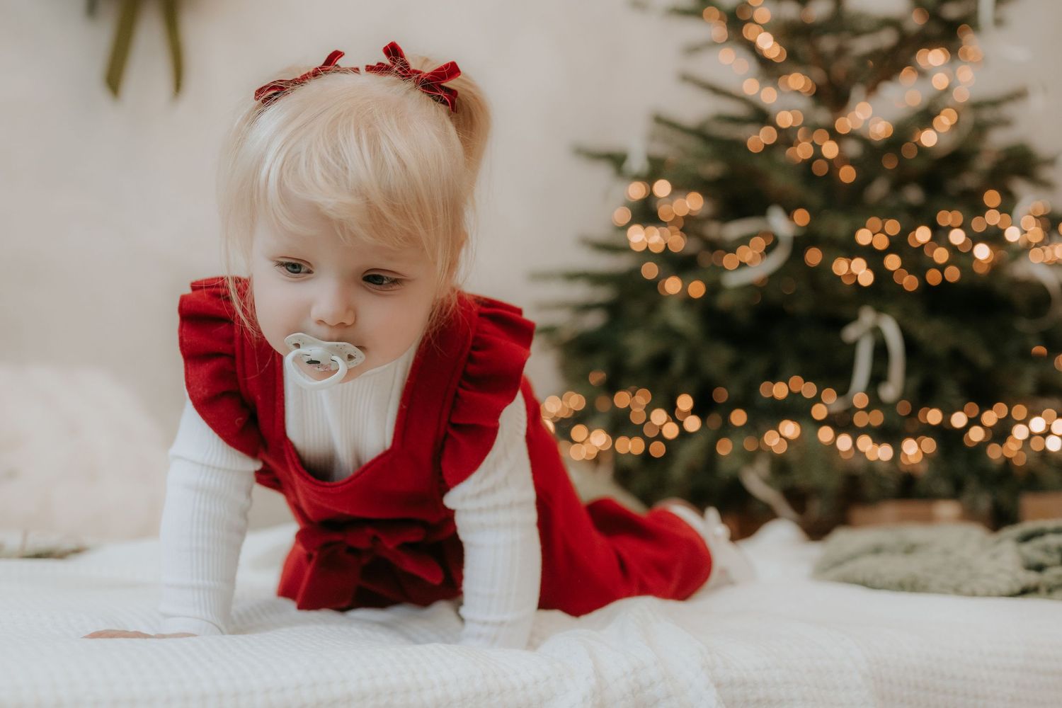 Blonde toddler girl in red ruffled Christmas dress with pacifier crawling near decorated holiday tree.