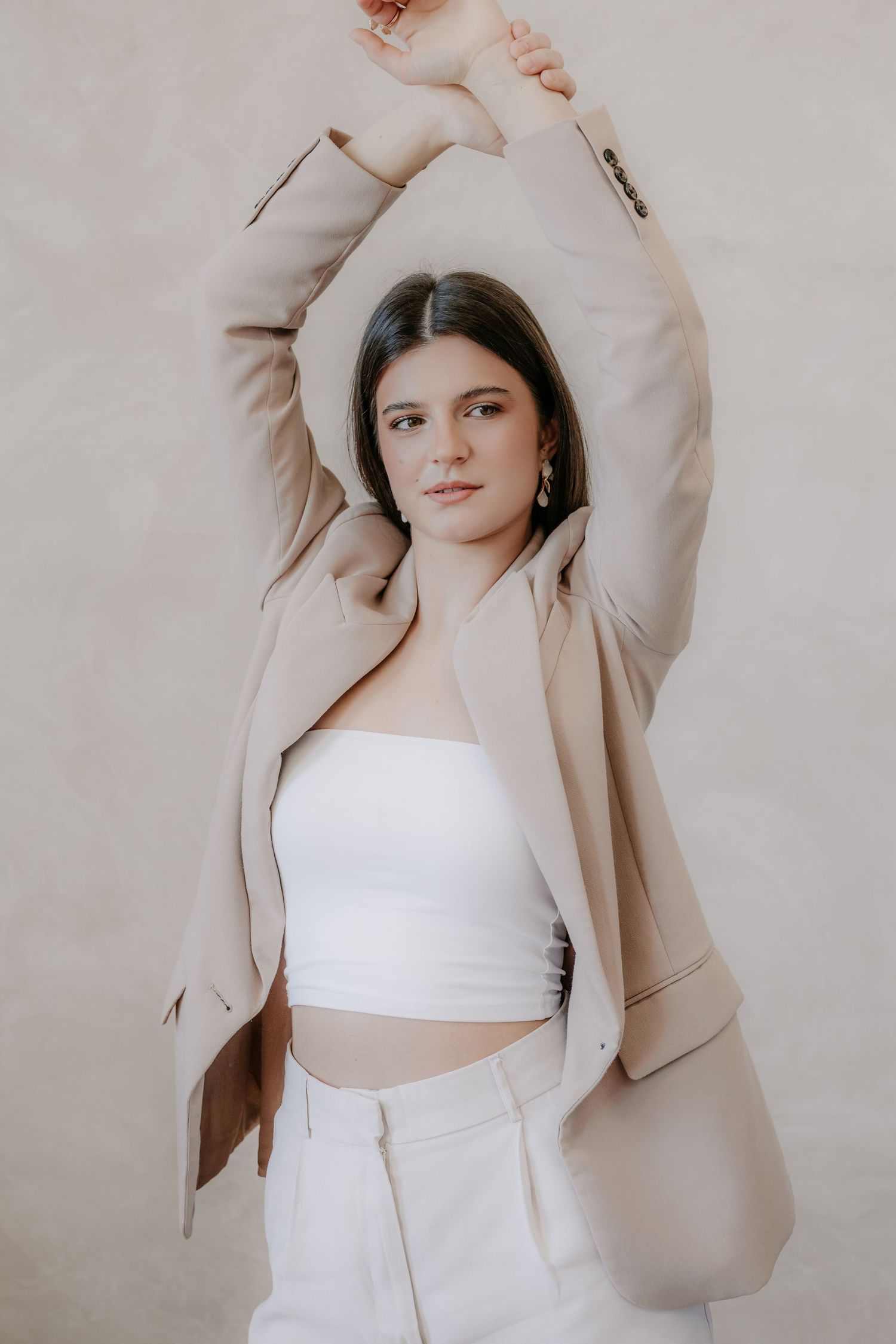 Young woman in beige blazer and white crop top poses with arms raised against neutral background.