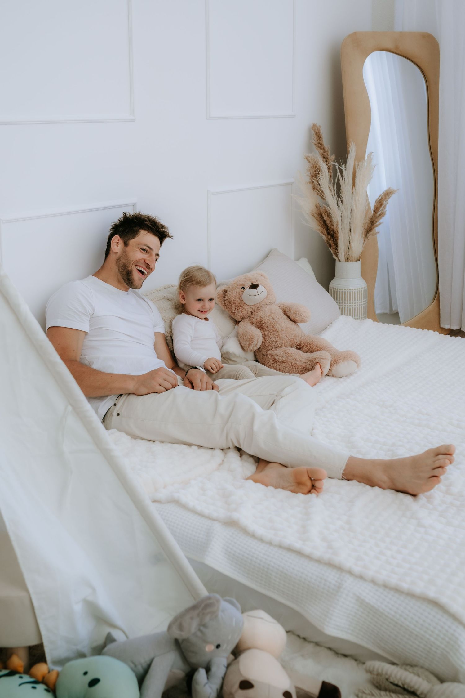 Father and toddler relaxing on a white bed with a teddy bear, surrounded by stuffed animals in a bright bedroom.