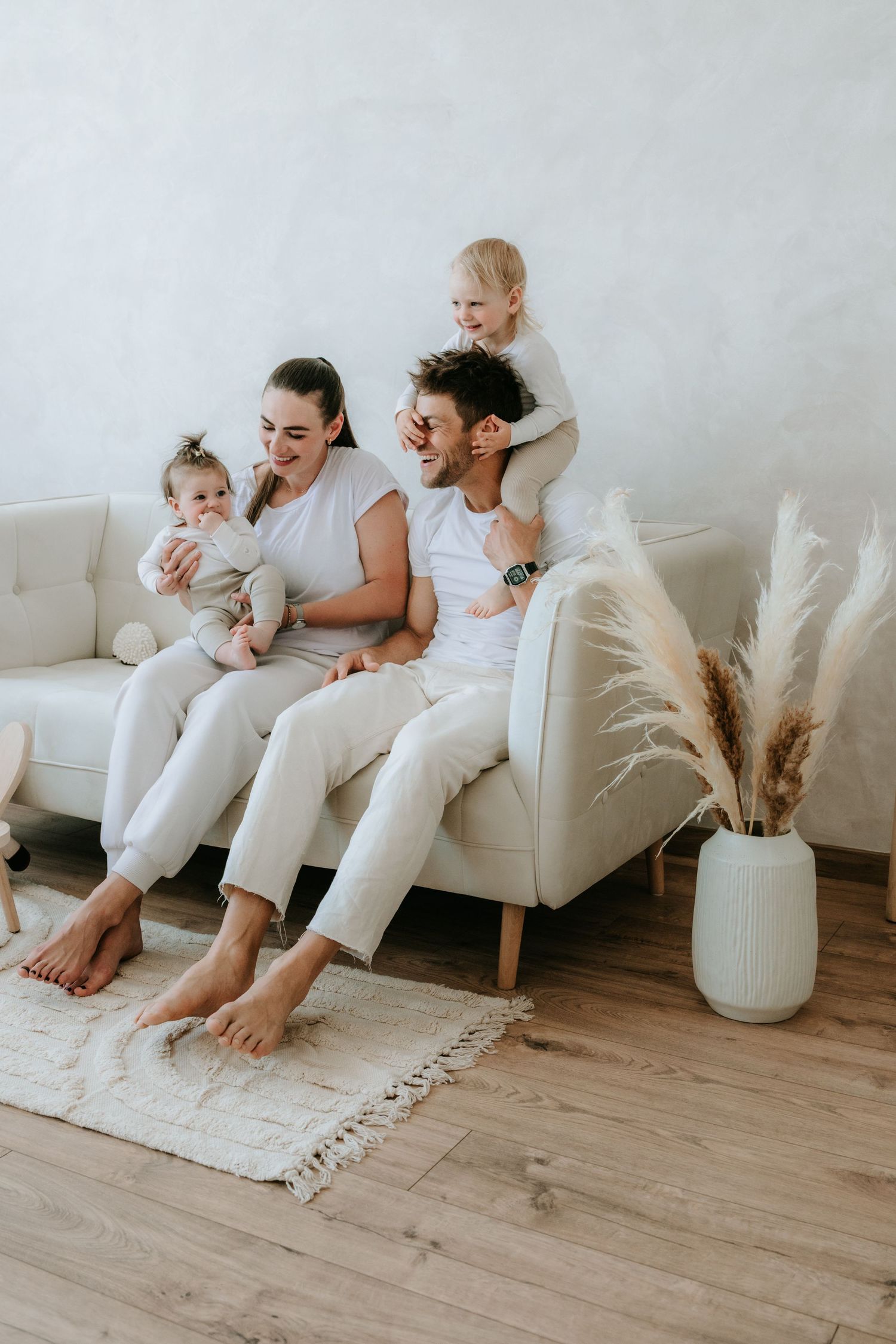 Happy young family with two toddlers sitting together on a white sofa in a bright modern living room.