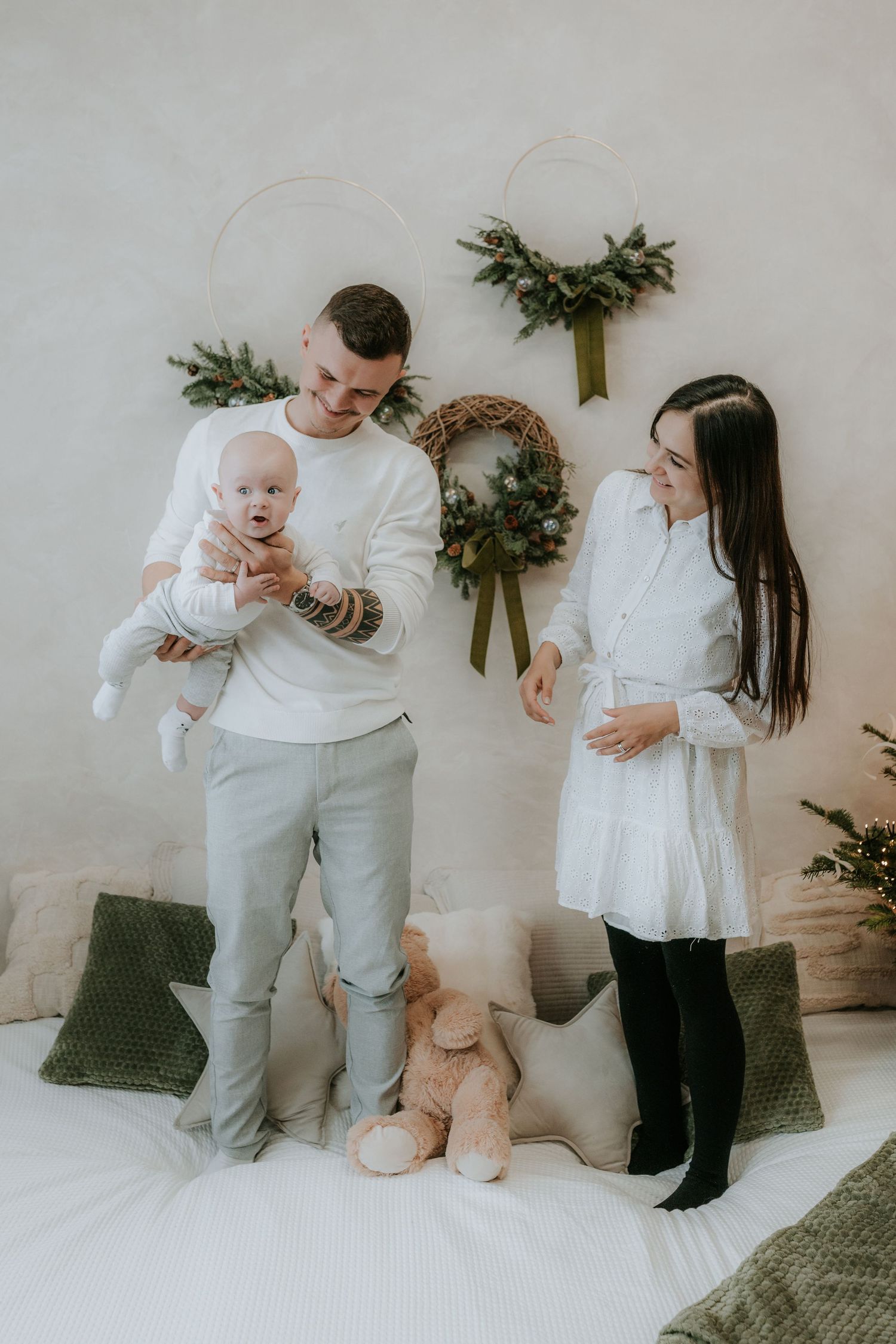 Young family with baby standing on bed with Christmas wreaths and holiday decorations in white room.