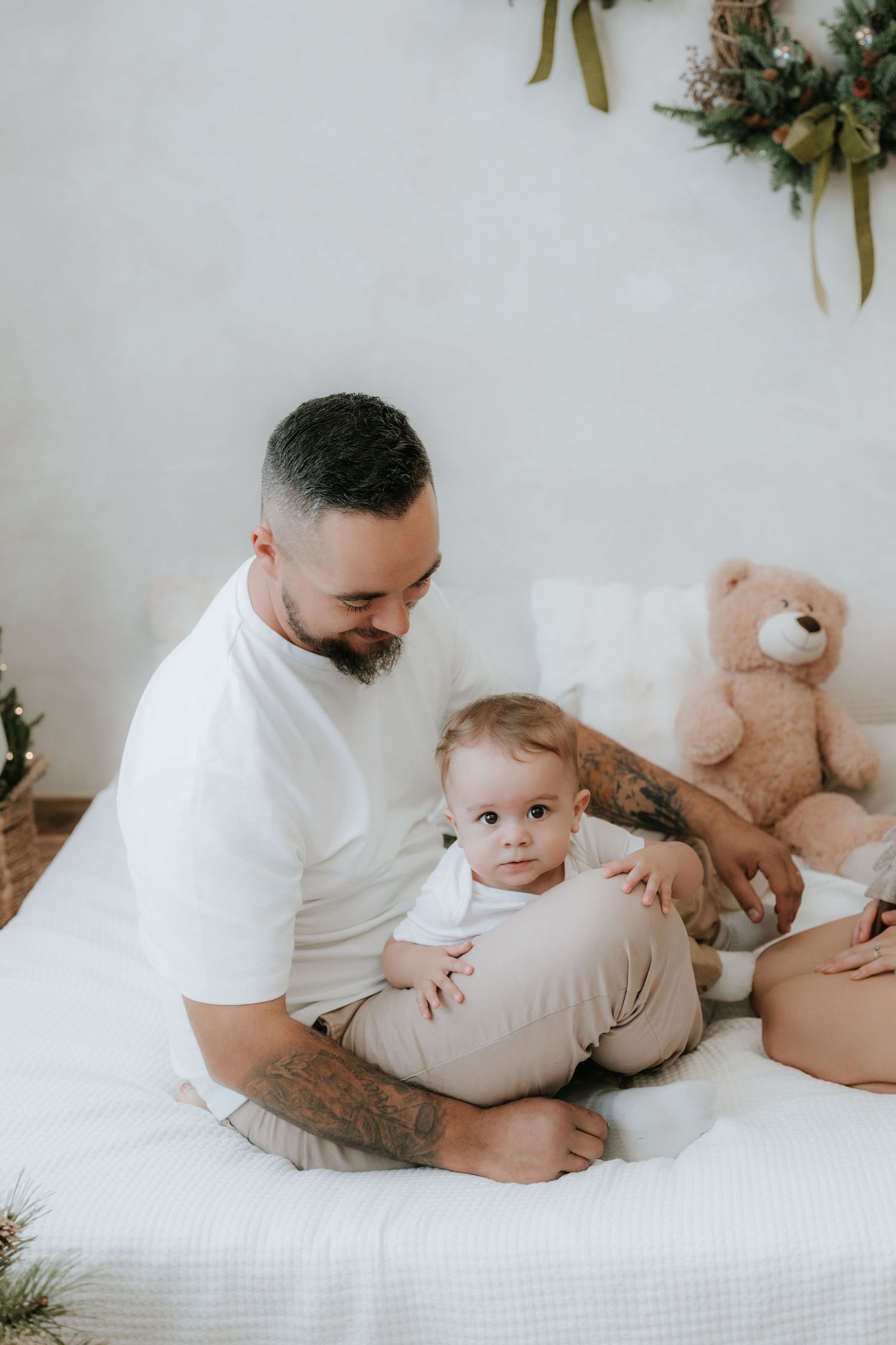 A tattooed father sits on a white bed holding his baby, with a teddy bear and holiday wreath decor in background.