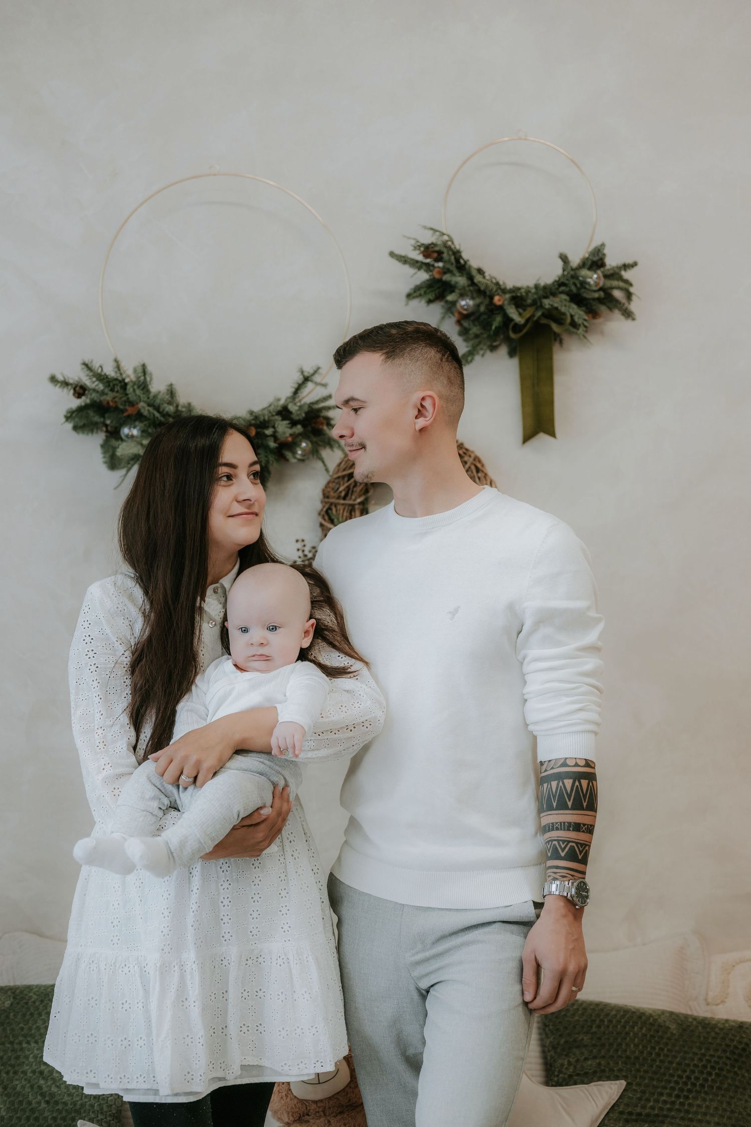 Young family with baby poses before holiday wreaths, wearing white outfits in a festive Christmas photo session.