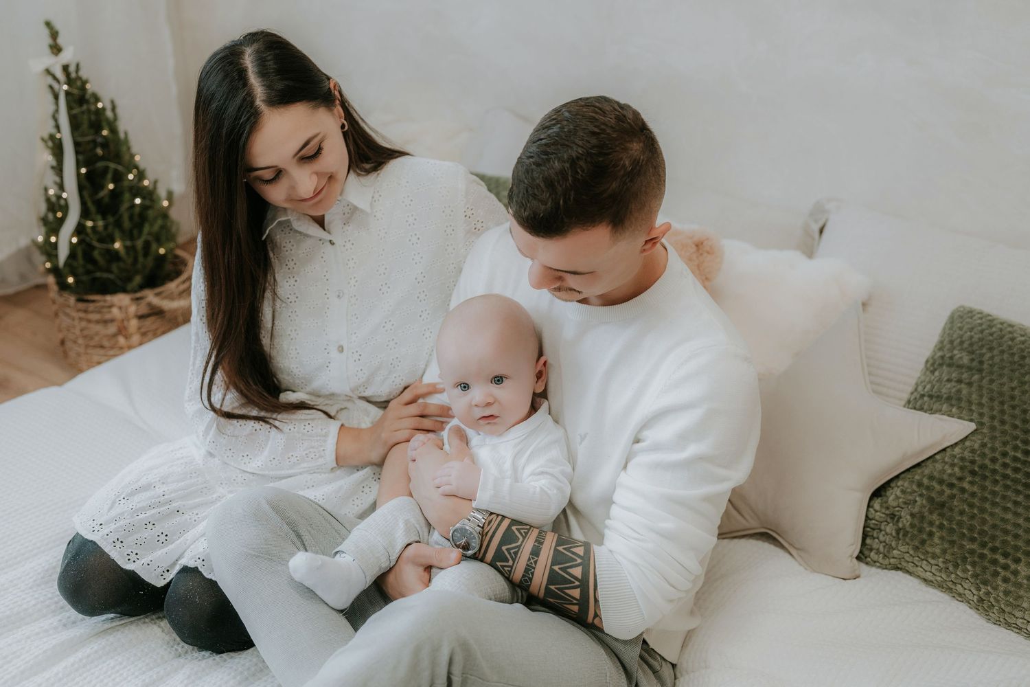 Young family in white outfits sitting on bed with newborn baby near Christmas tree in bright cozy room.