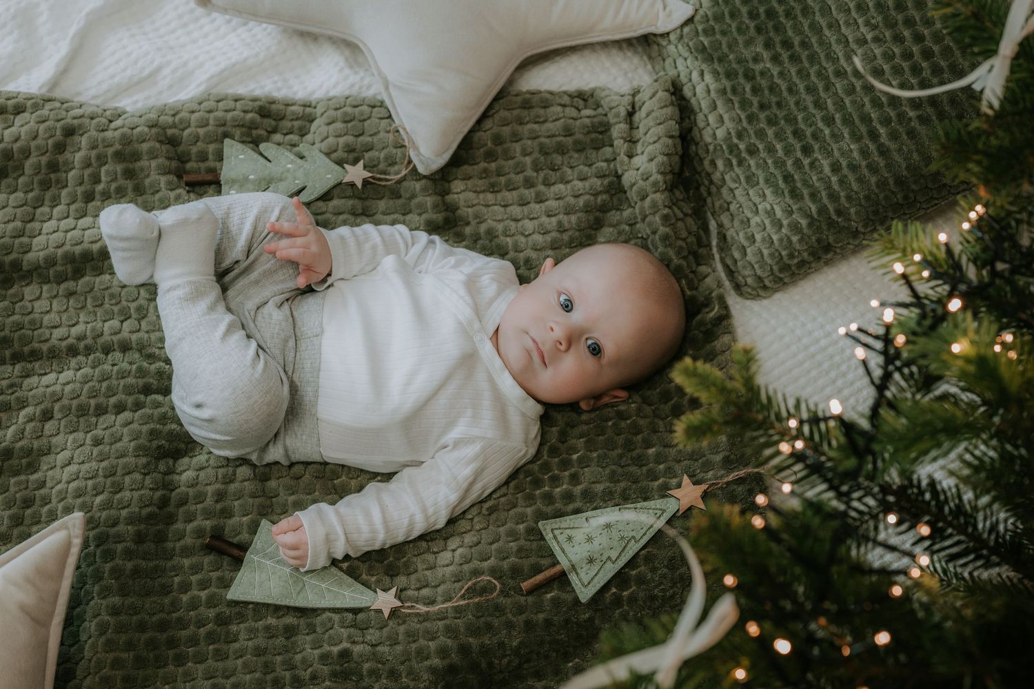 Baby in white onesie lying on green blanket beside decorated Christmas tree with fairy lights and festive ornaments.