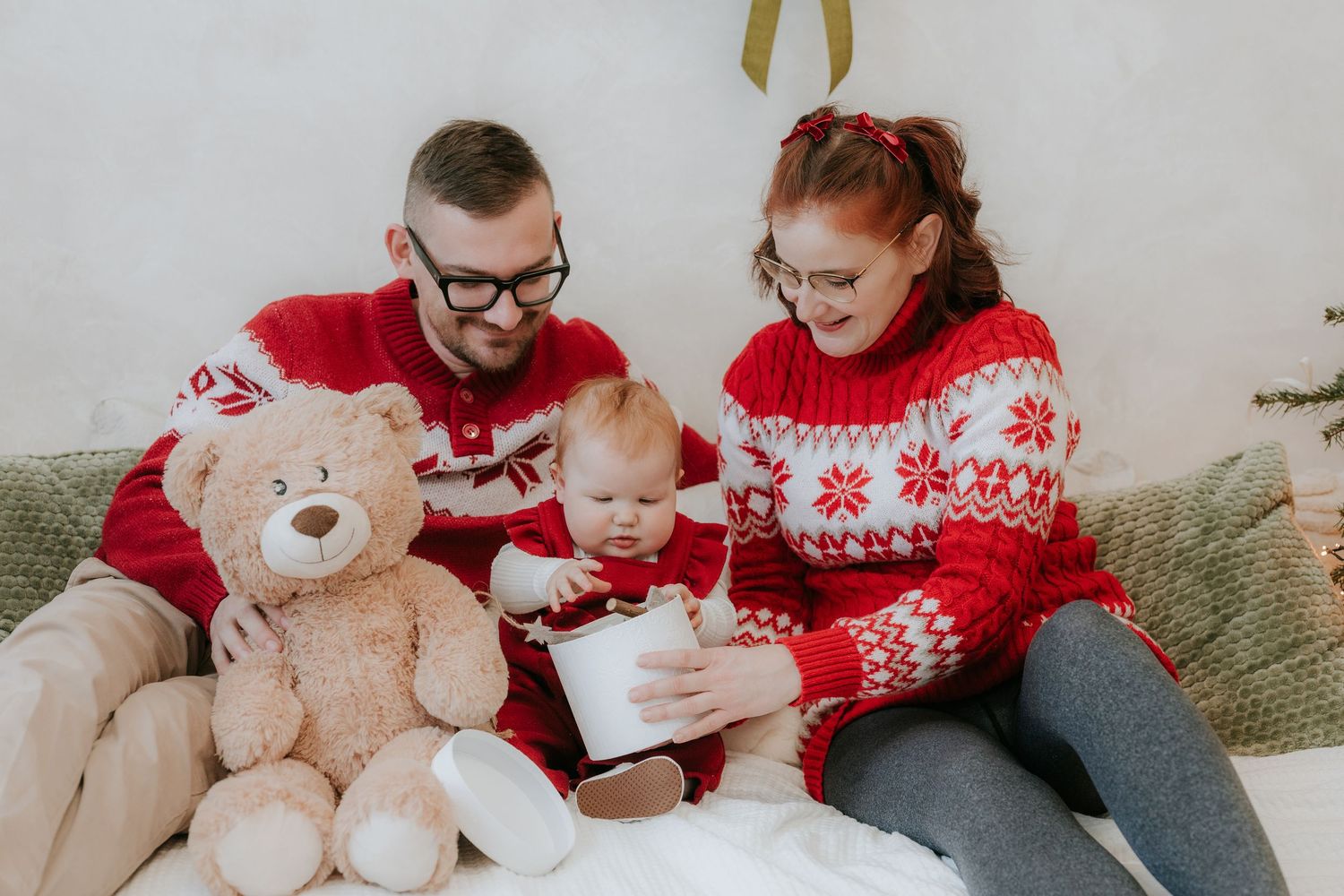 Family in matching red Christmas sweaters sitting on couch with teddy bear, sharing holiday cup with baby.
