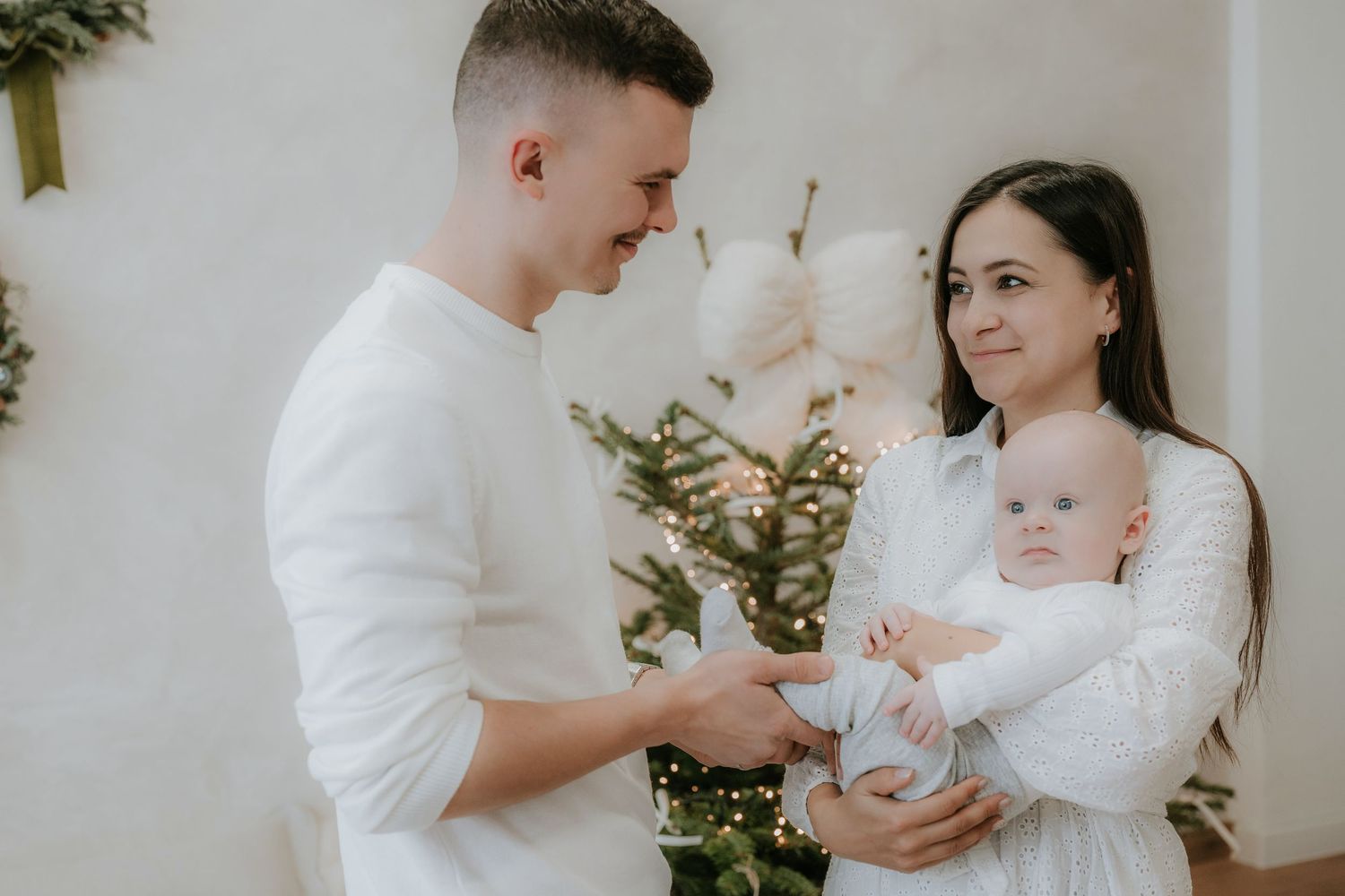 Young family in white outfits celebrating Christmas, parents smiling at newborn baby near decorated tree.