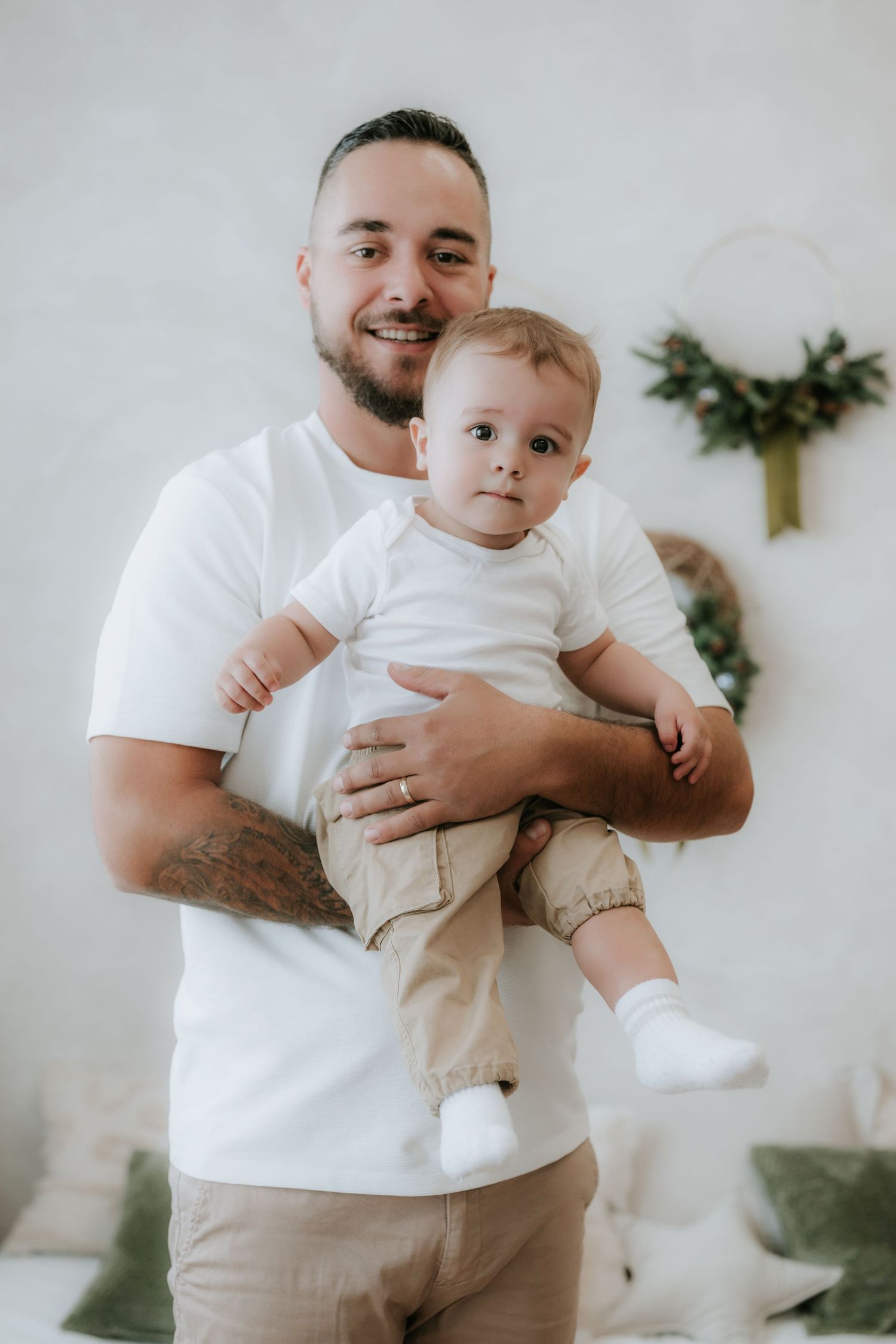 Smiling father with tattoos holding his baby boy, both wearing white shirts and khaki pants, in a cozy decorated room.