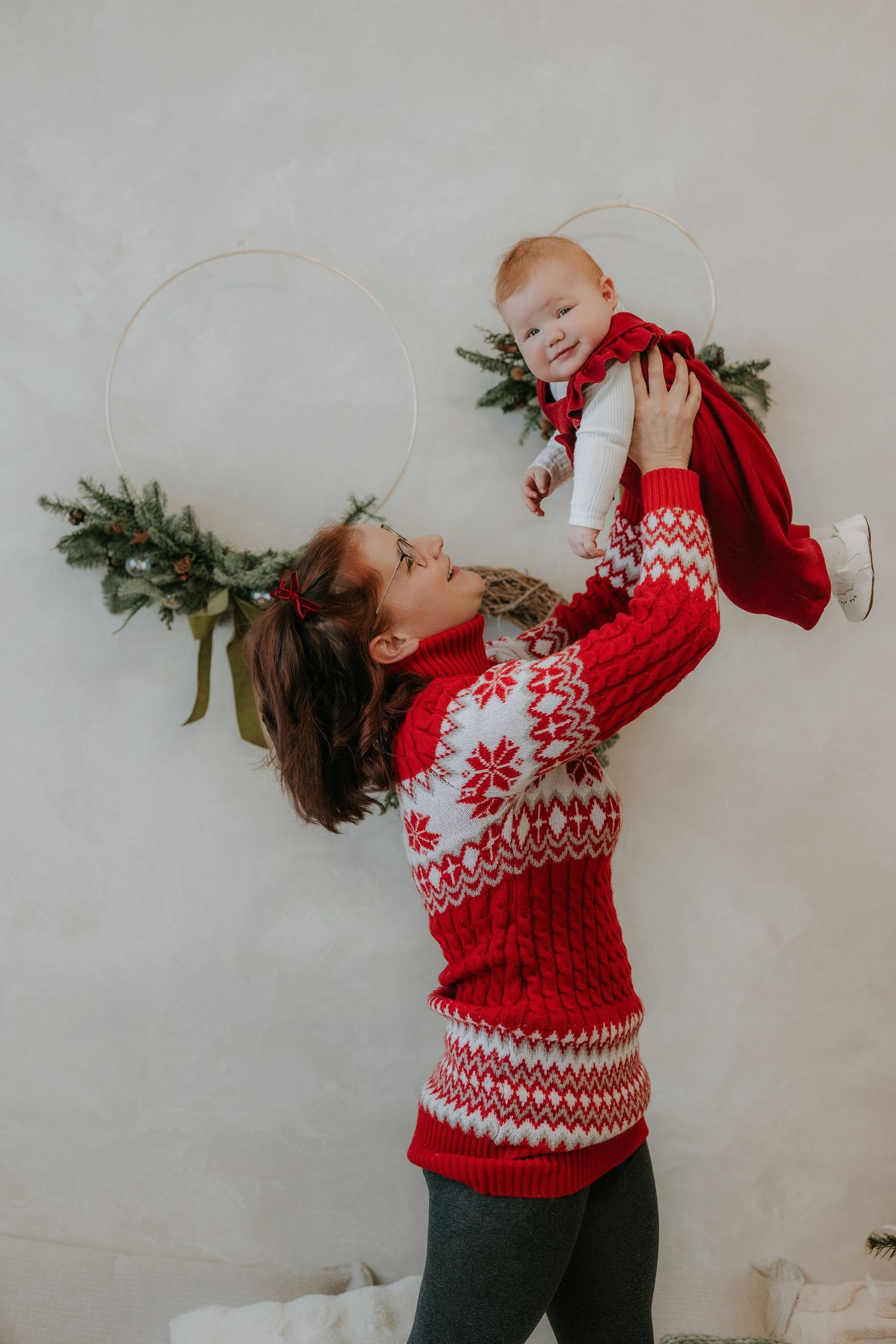 Mother in red Nordic sweater joyfully lifts her baby dressed in red during a festive Christmas photo session.