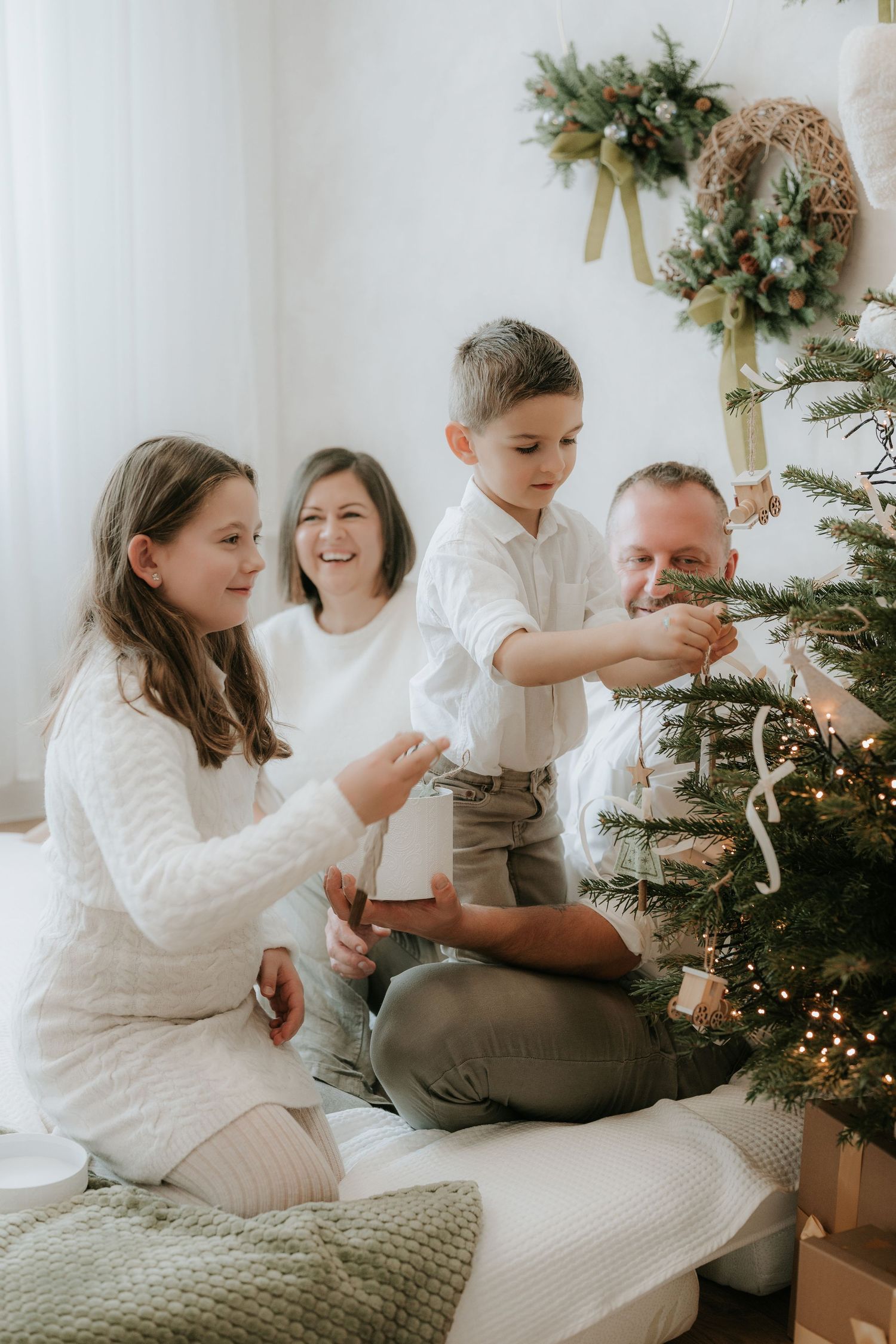 Family of four decorating a Christmas tree together in a bright white room with holiday wreath decor.