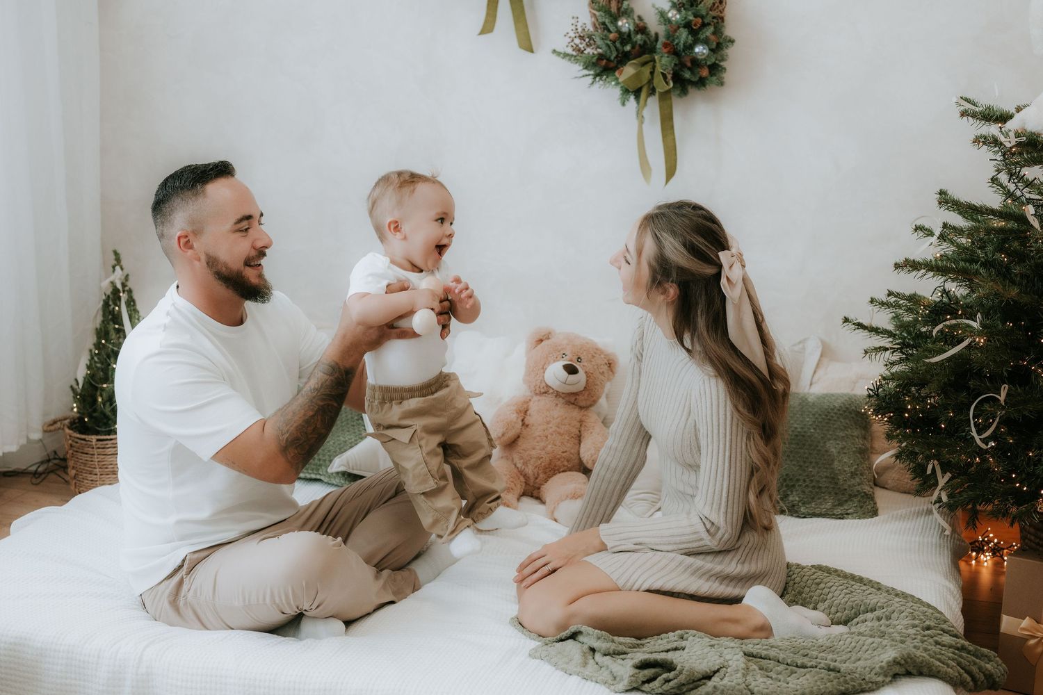 A happy young family sits on a bed near a Christmas tree, with parents playing with their baby and a teddy bear.