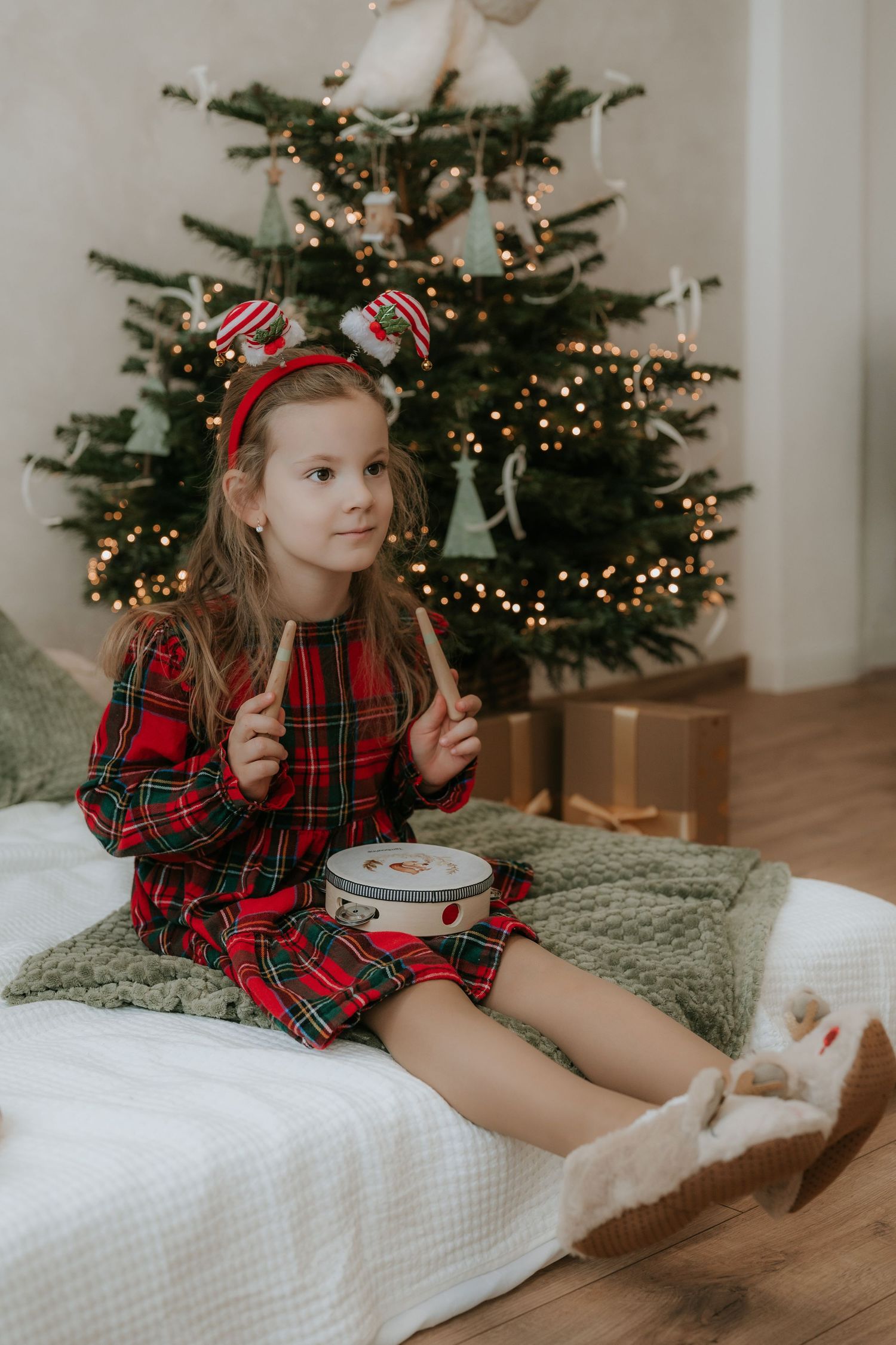 Girl in red plaid dress sits by decorated Christmas tree holding drumsticks with a cookie tin, wearing festive reindeer headband.