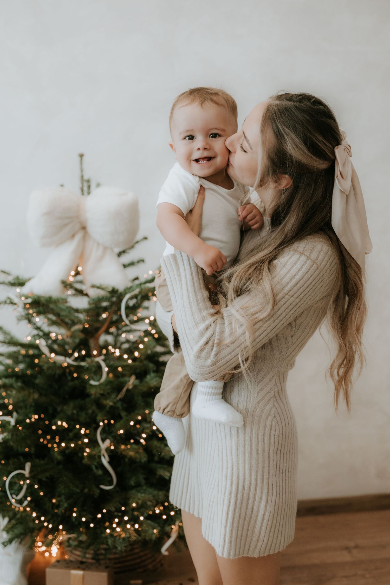 Young mother kissing happy baby near decorated Christmas tree with warm fairy lights.