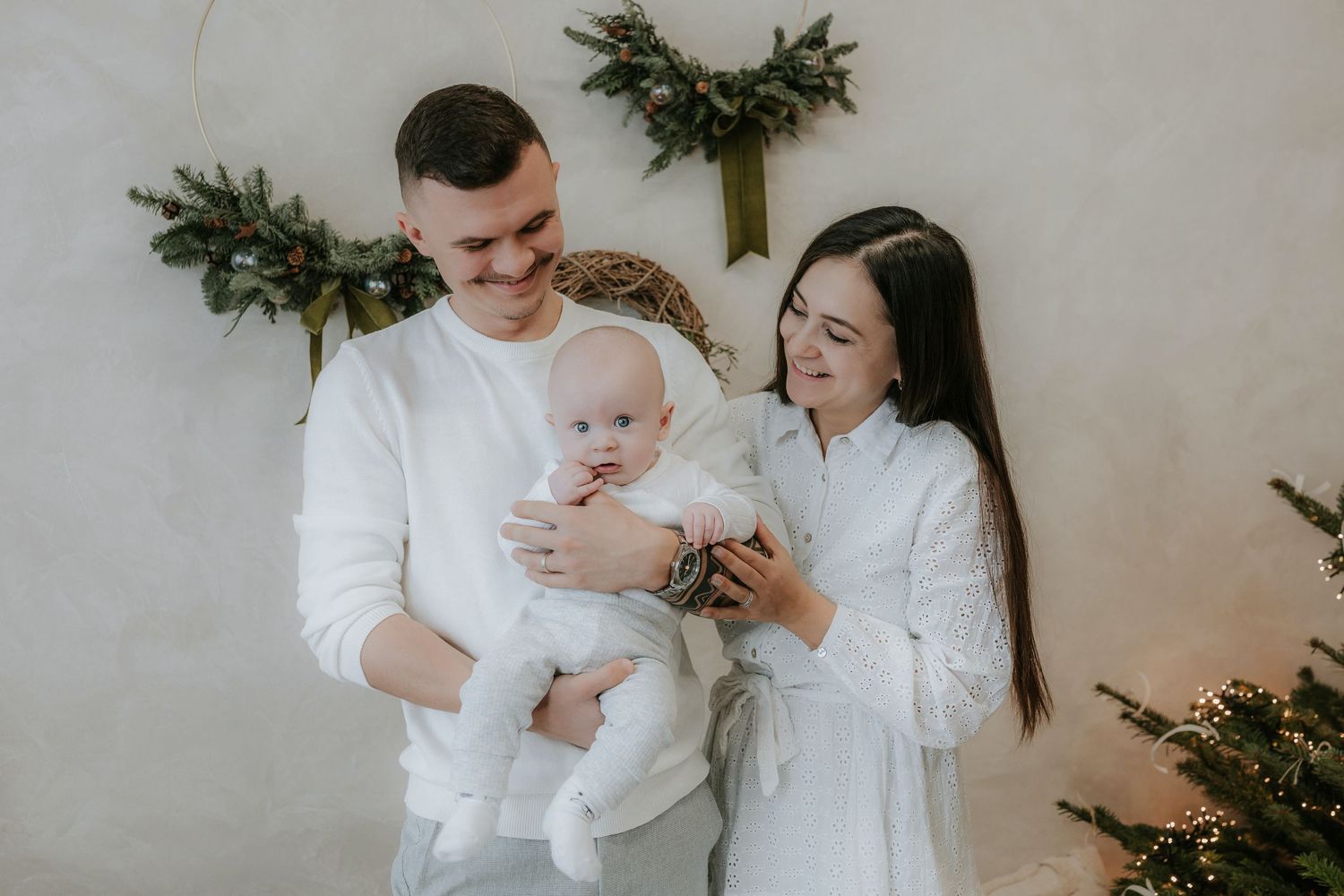 Young family in white outfits celebrating Christmas, parents holding baby near decorated tree and wall.