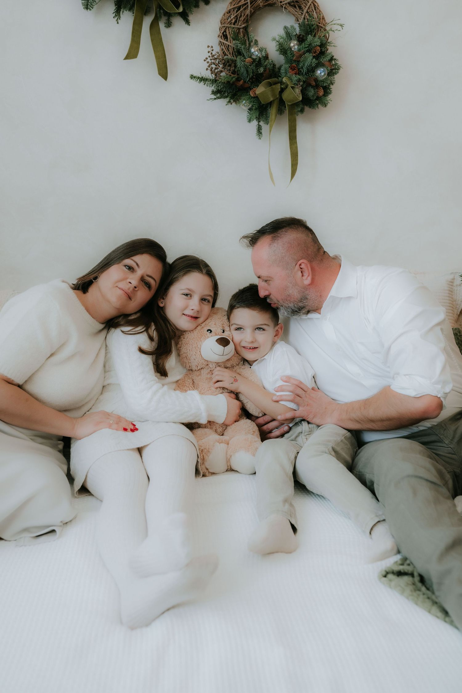 Family of four in white outfits cuddles on bed with teddy bear under Christmas wreath.