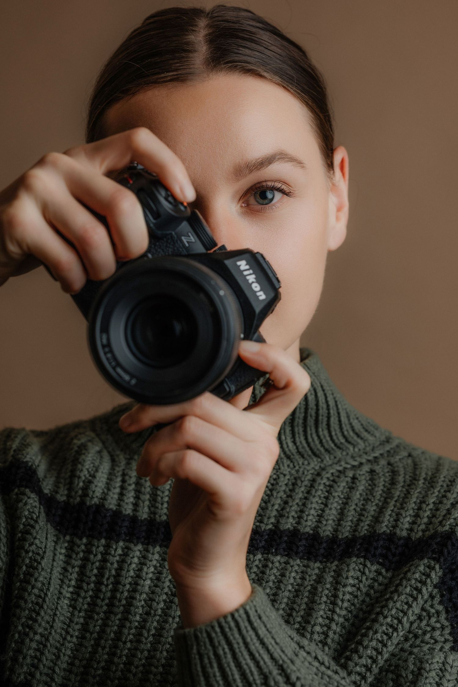 Young woman in green knit sweater holding a Nikon DSLR camera up to her eye, photographing against a beige background.