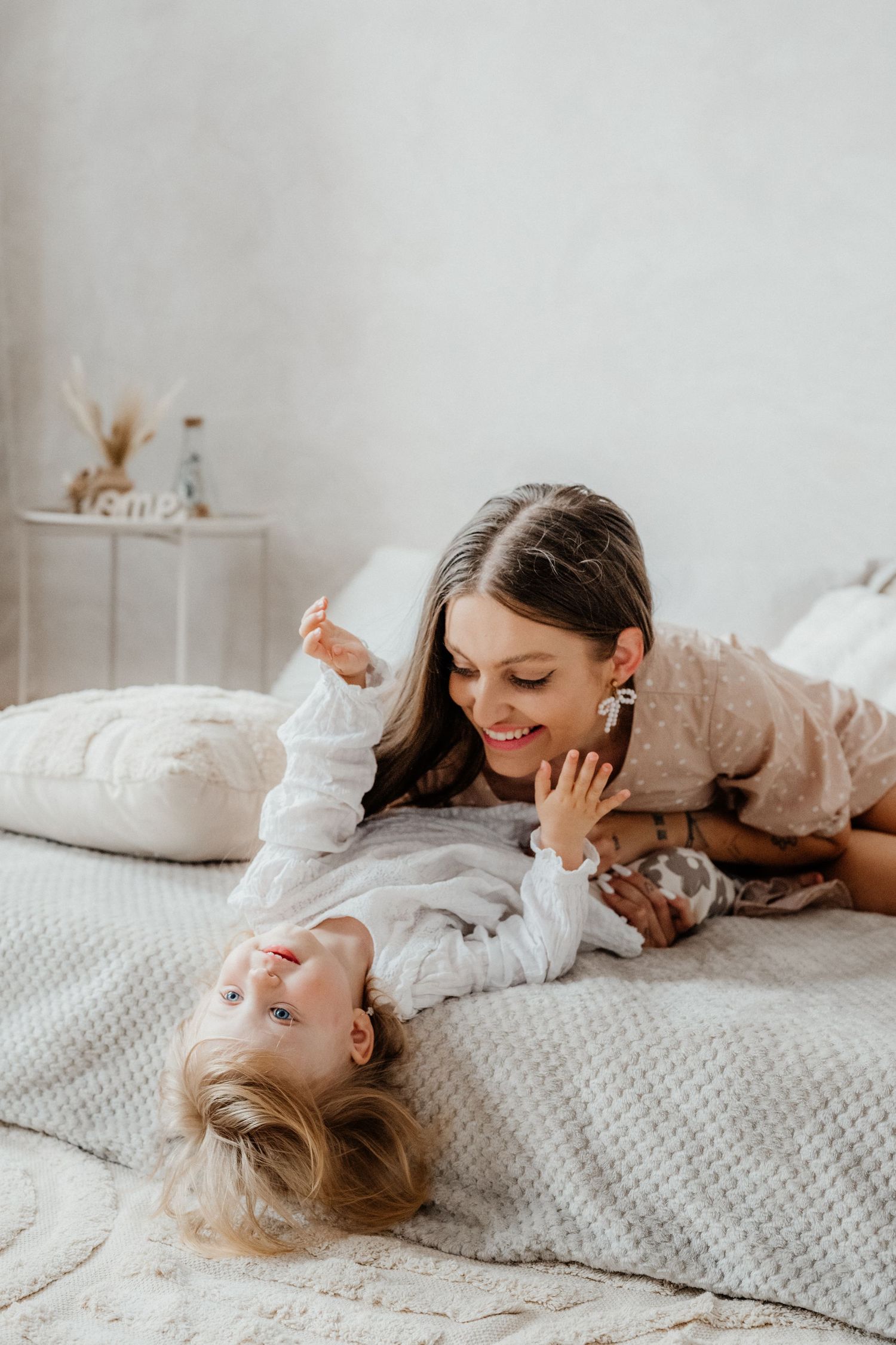 A smiling mother lying on a bed playing with her young toddler child in a bright white bedroom.