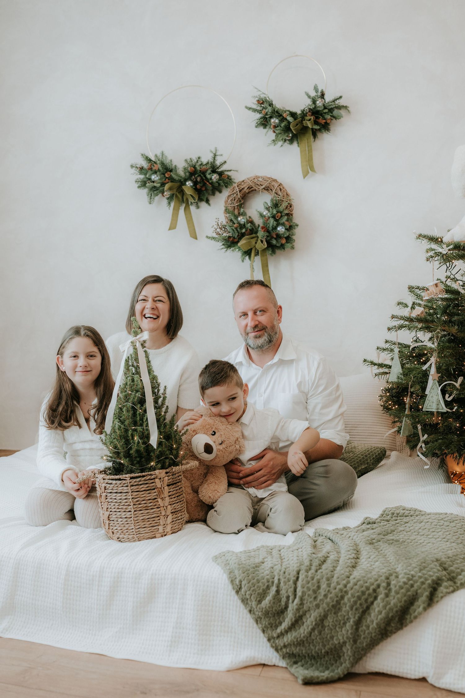 Family of four sitting together by a Christmas tree with wreaths on wall, smiling and holding teddy bear and mini tree.