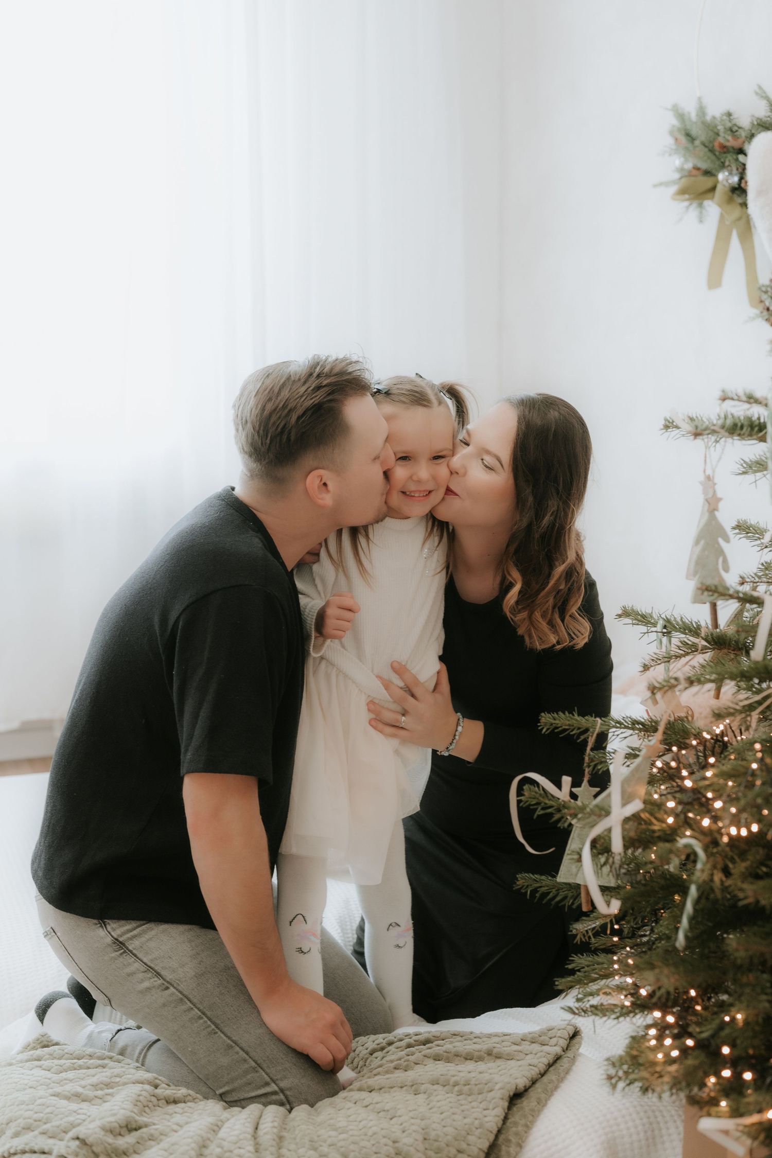 Parents kissing their young daughter on the cheeks beside a decorated Christmas tree in a bright white room.
