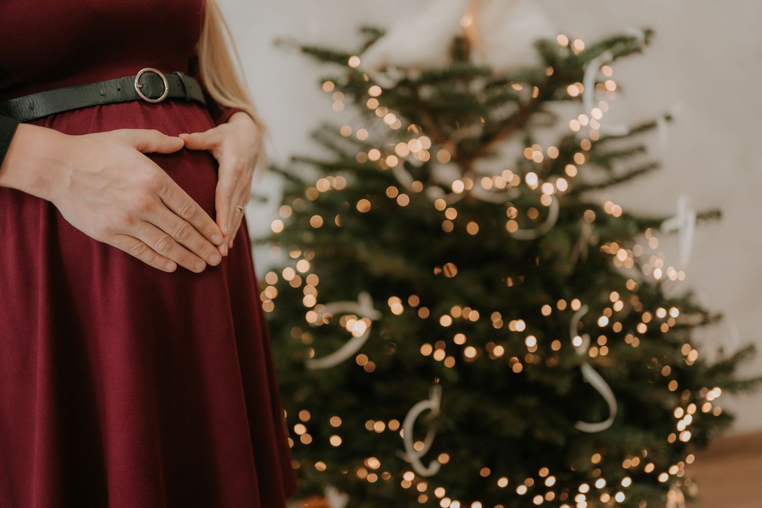 Pregnant woman in burgundy dress cradling baby bump in front of a decorated Christmas tree with bokeh lights.