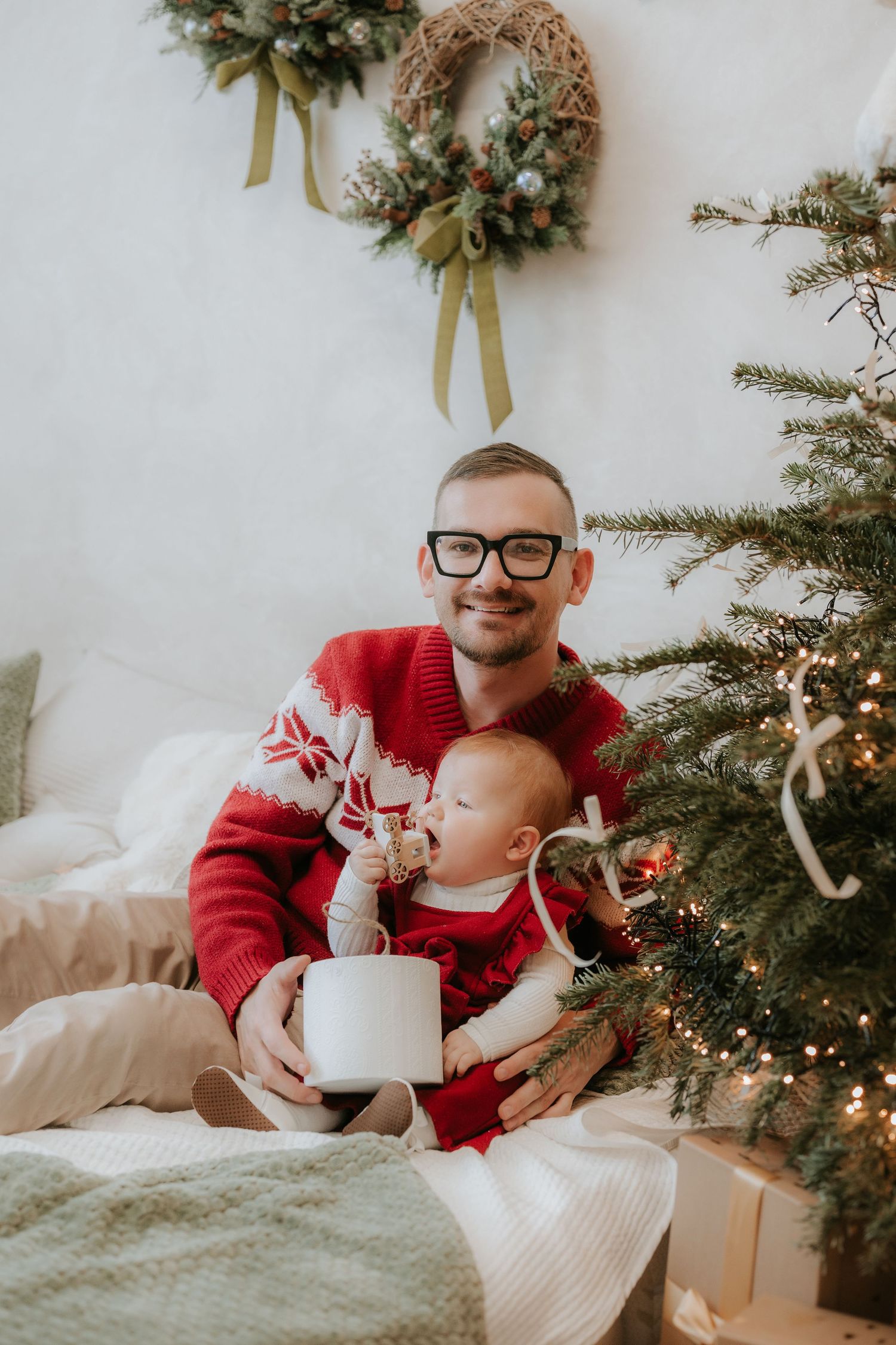 Father in red Christmas sweater holds baby near decorated tree with wreath and gifts in cozy holiday setting.