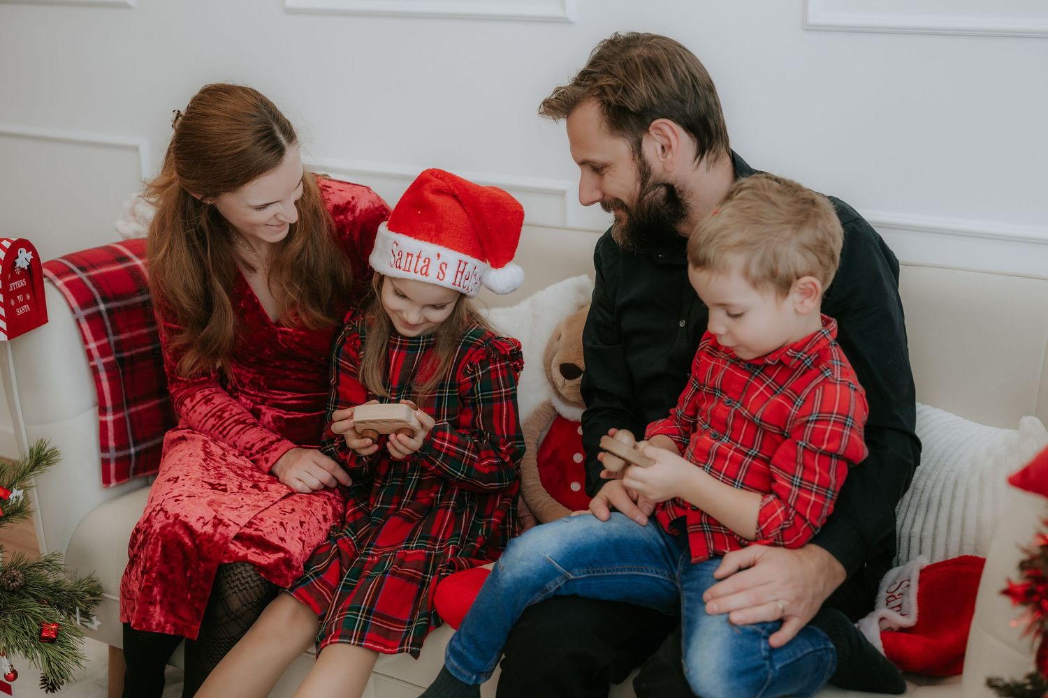 Family of four in festive red plaid outfits sitting on a white couch, sharing holiday gifts together.